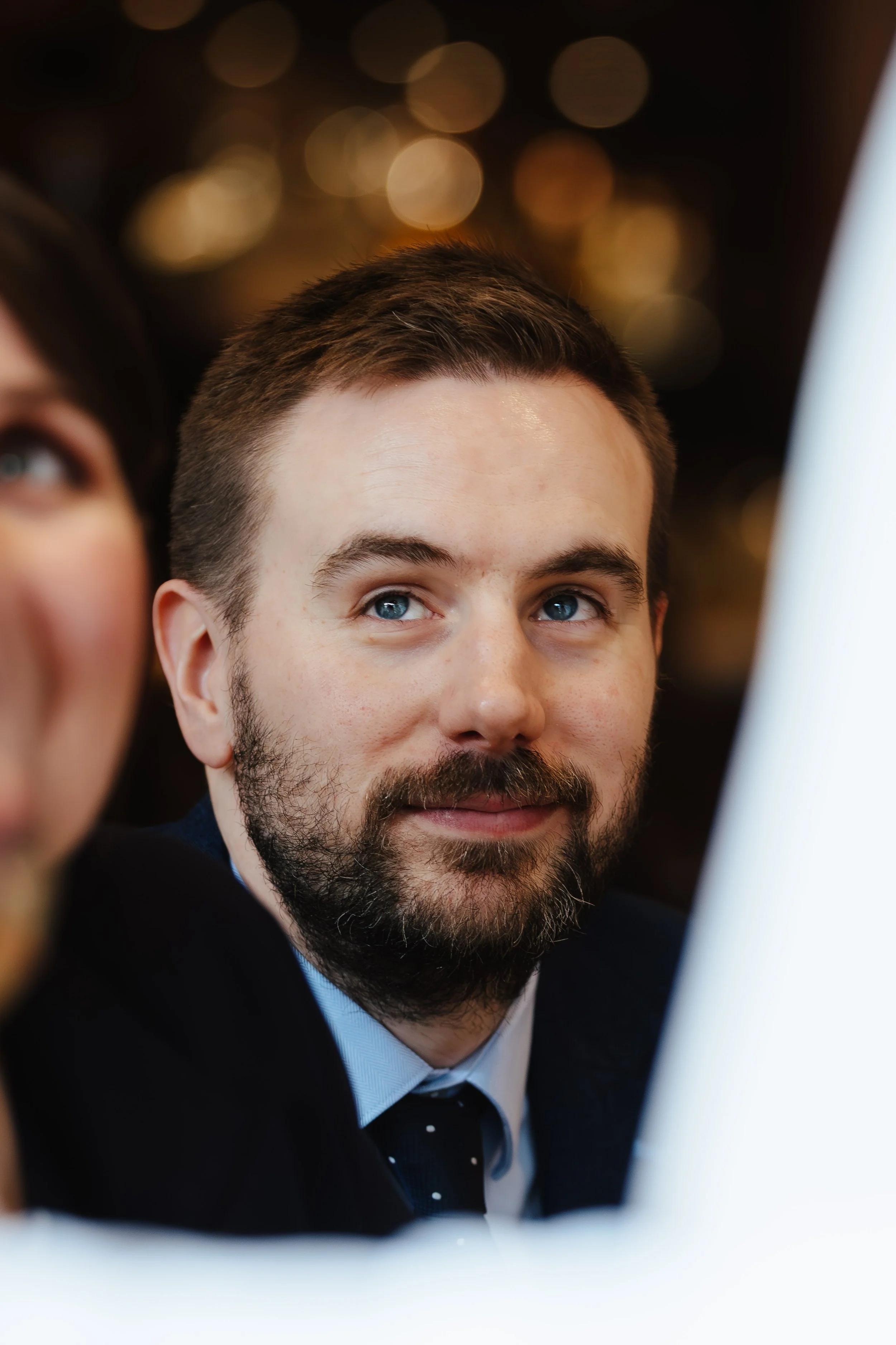 A man with a beard and blue eyes looking at a computer screen, with warm, bokeh lights in the background.