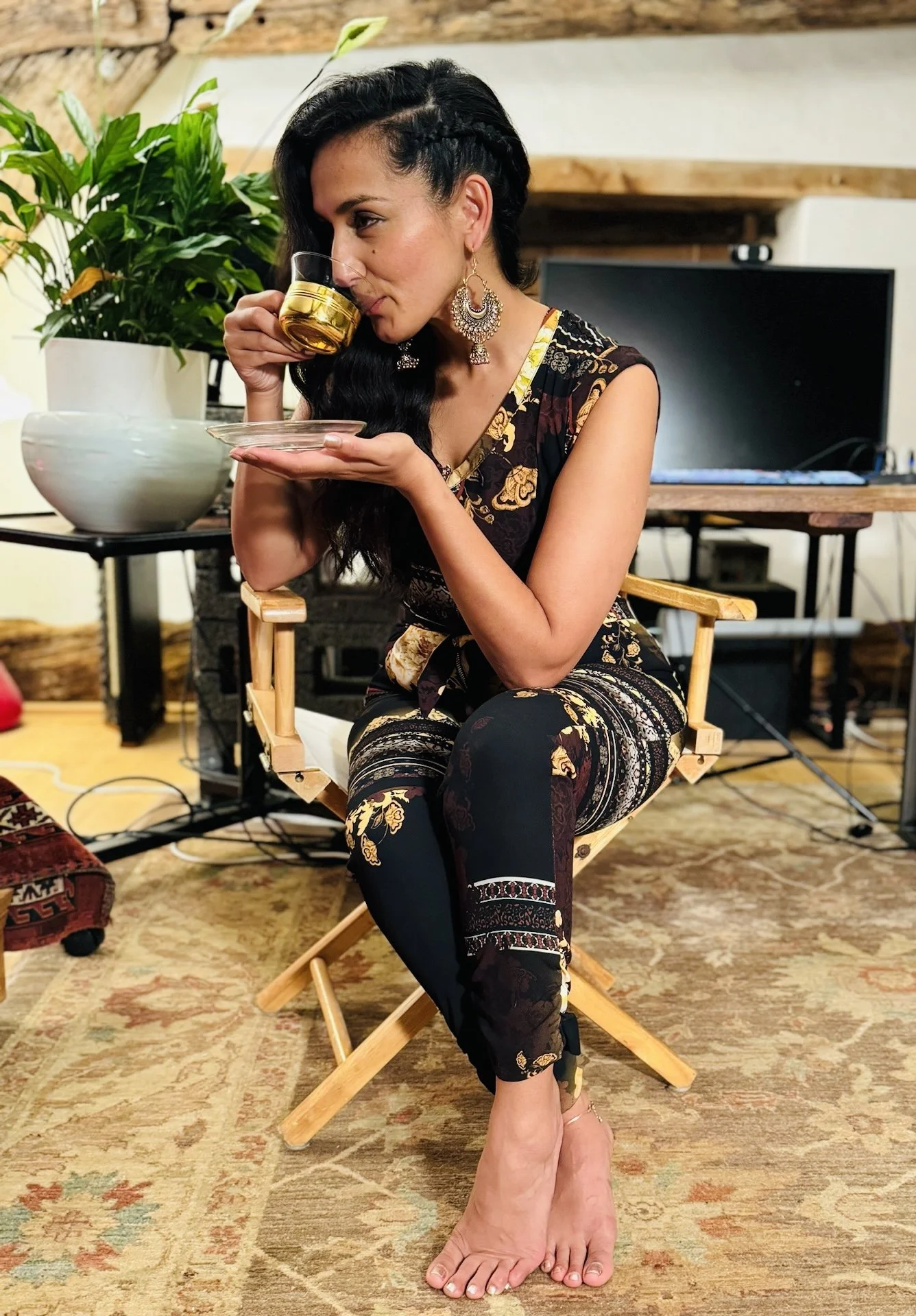 Woman with dark hair styled in waves and a braid, wearing large earrings and a patterned, sleeveless dress, sitting on a wooden chair, drinking from a glass cup with a gold base, with a large plant and a television in the background.