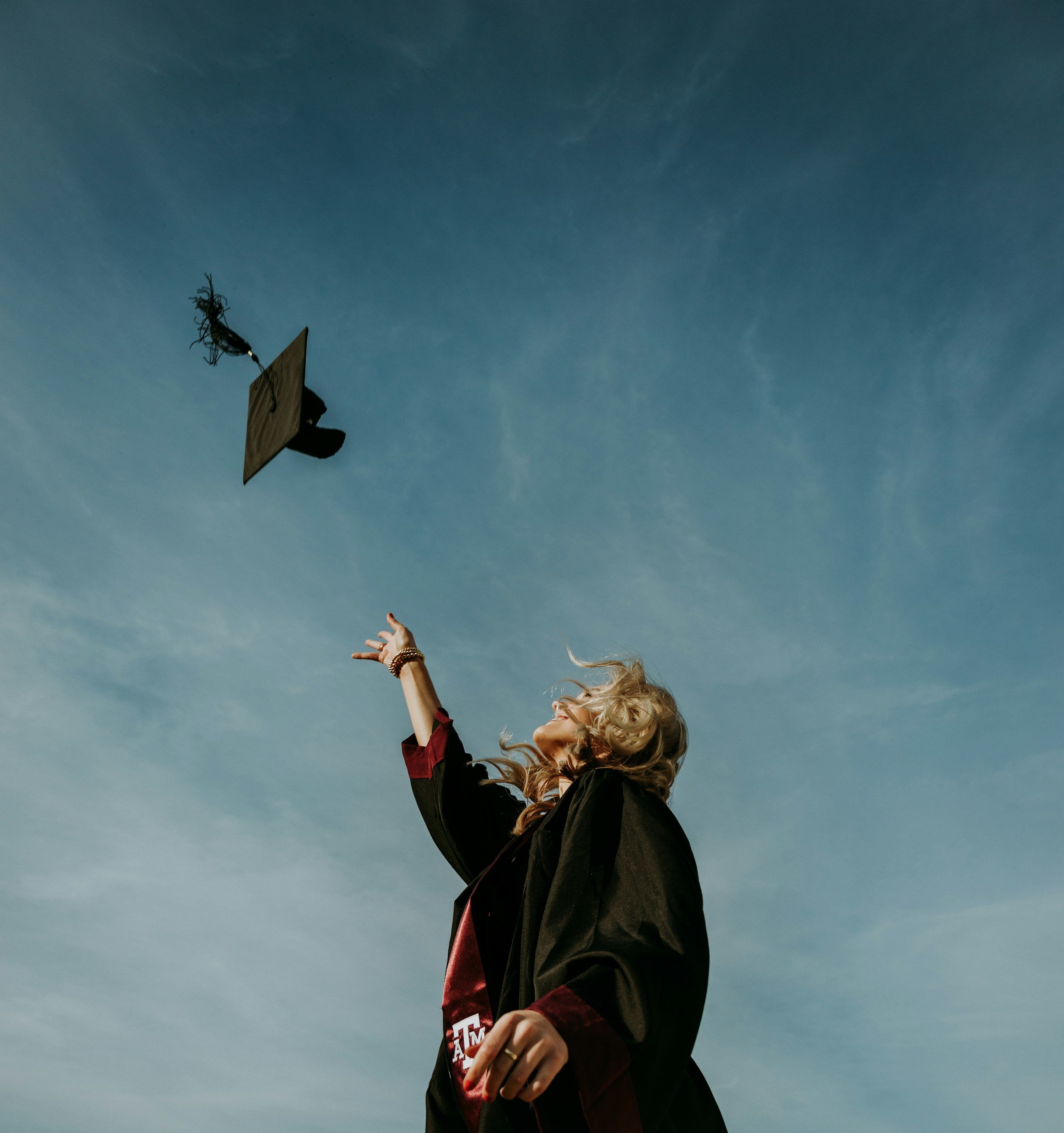 Young woman outside throwing graduation cap into the blue sky. She's wearing a black graduation gown with red details and smiling upwards