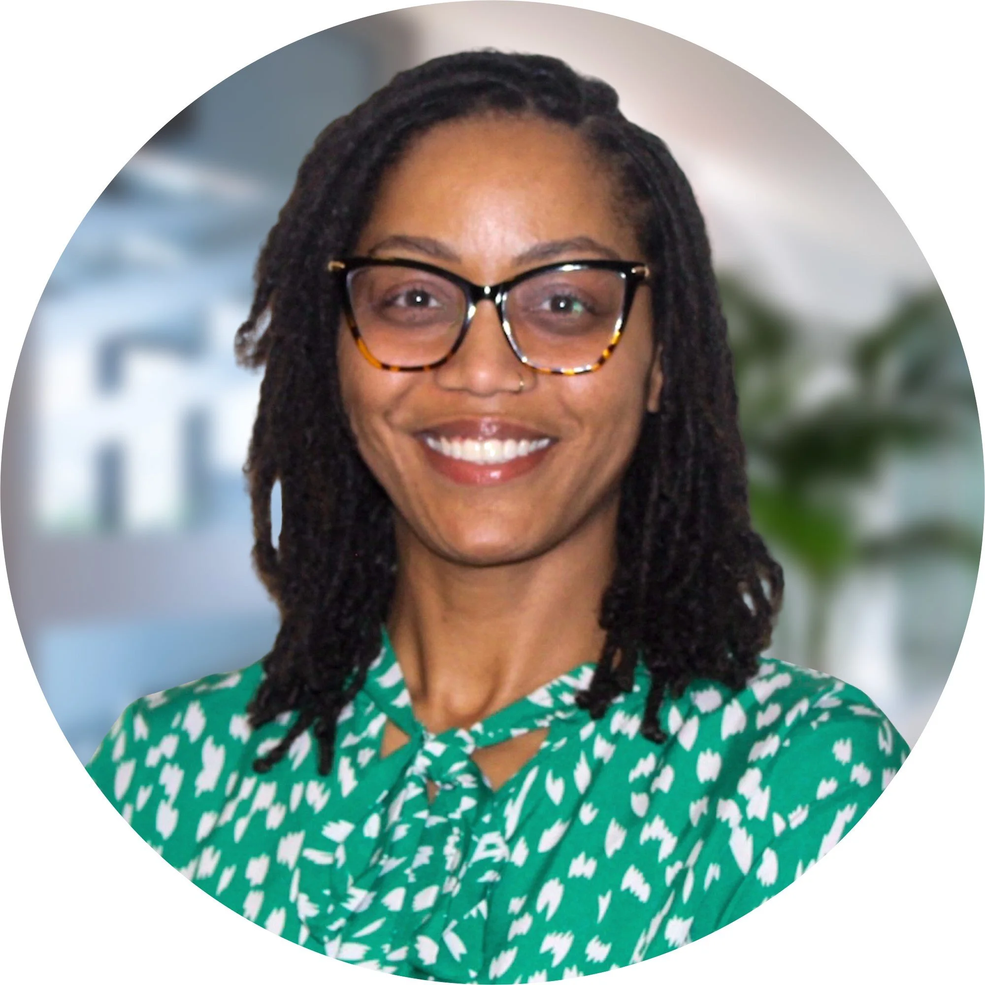 A woman with shoulder-length dreadlocks, wearing black and tortoiseshell glasses, smiling, in a green and white patterned blouse, with a blurred office background and a potted plant.