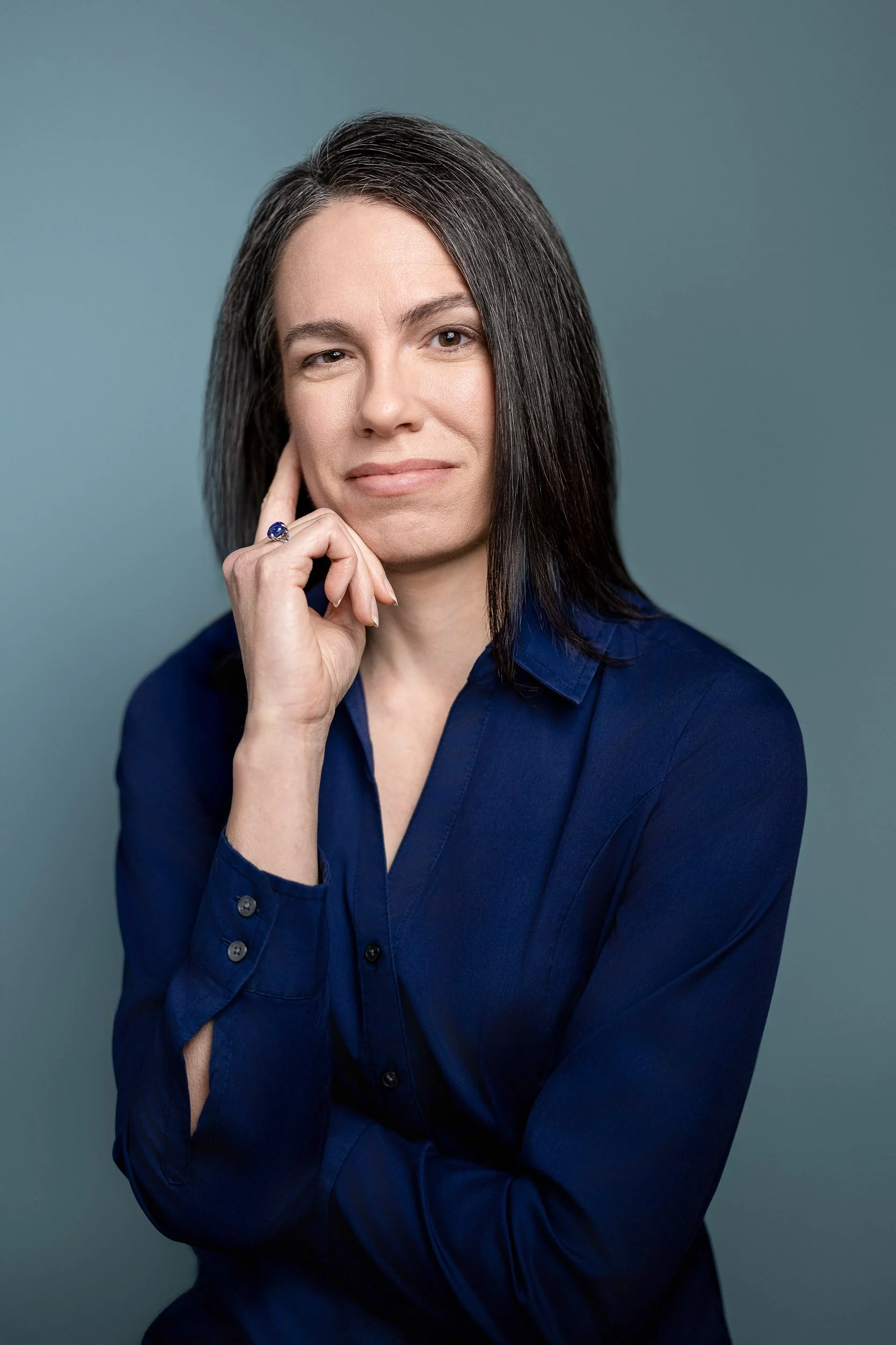 A woman with shoulder-length dark hair in a navy blue shirt, posing with her hand near her chin against a plain blue background.
