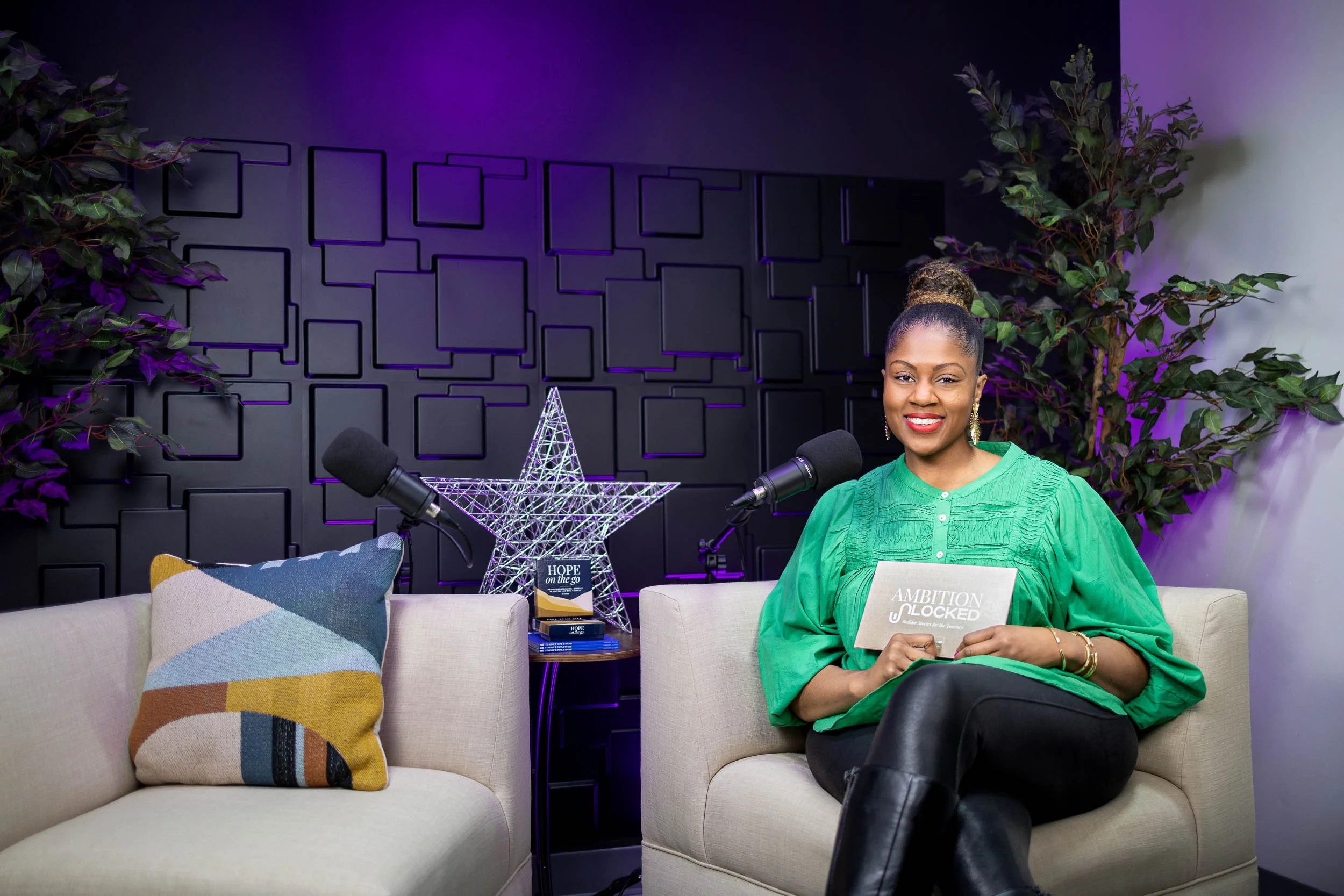 A woman smiling, sitting on a beige sofa with a colorful cushion, holding a book titled "Ambition Unlocked" in a brightly lit room decorated with purple and black accents, microphones on stands nearby, and a decorative star and plants in the backgrou