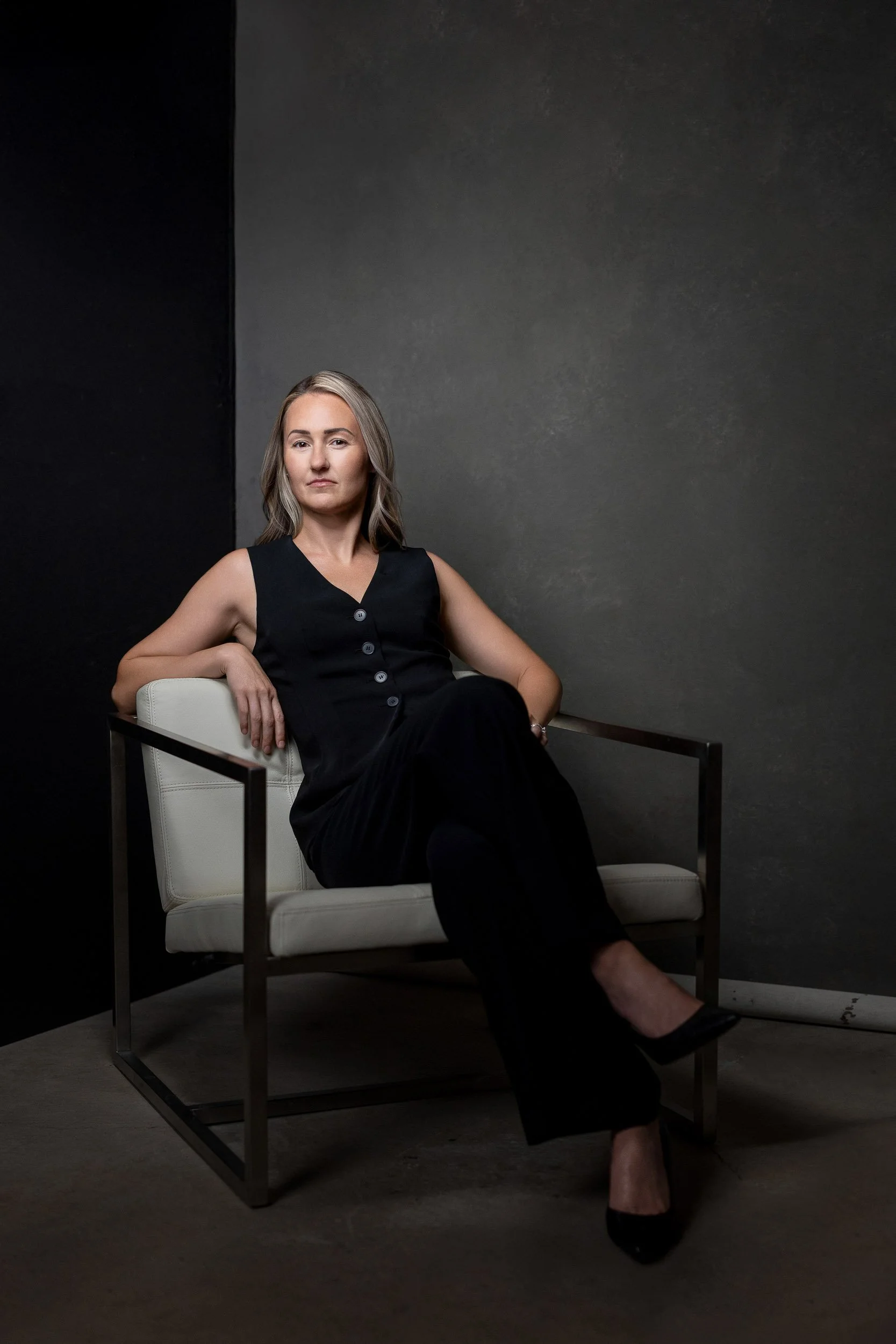 A woman in a sleeveless black dress sitting on a white chair with metal frame against a dark textured background.