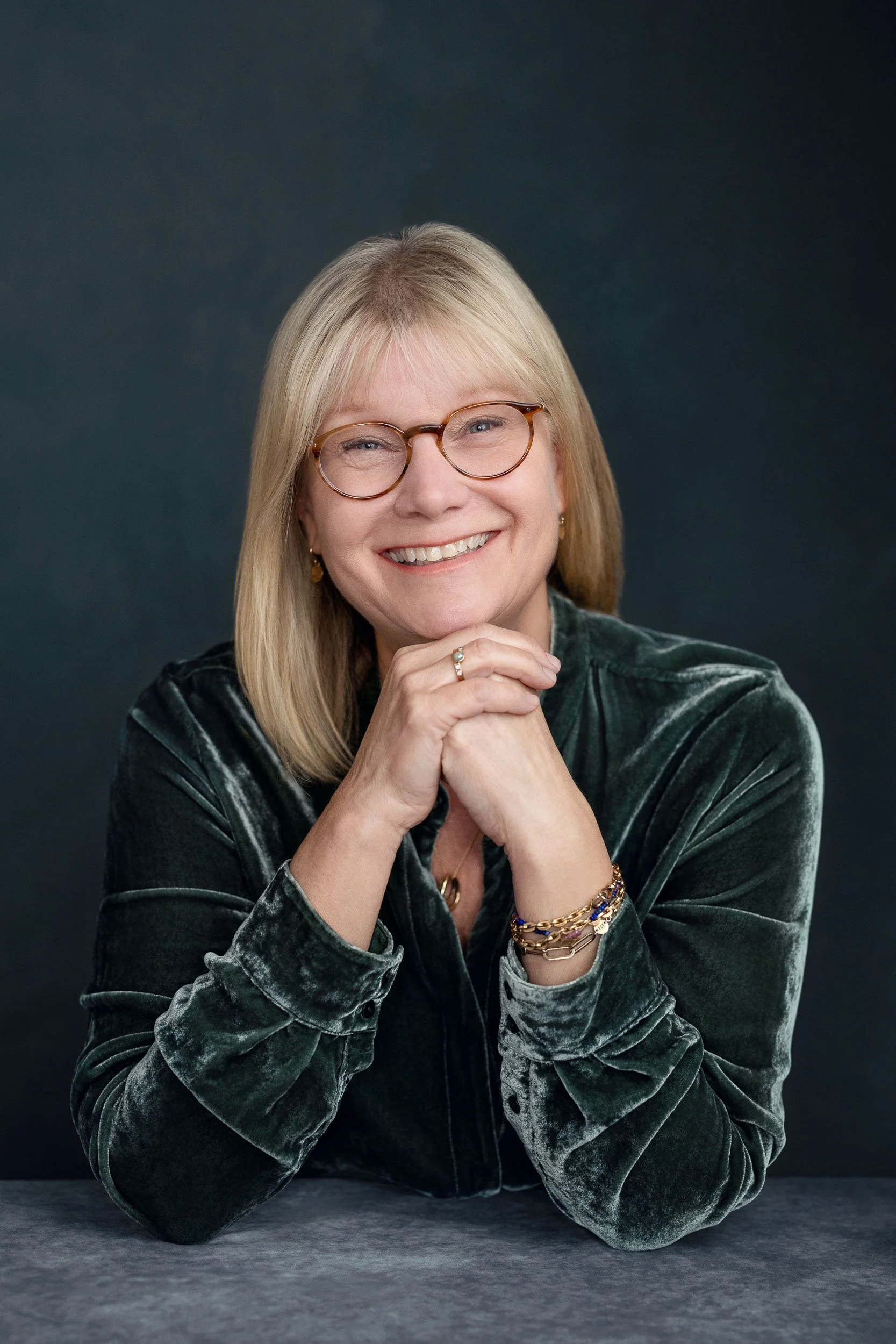 Portrait of a smiling middle-aged woman with blonde hair, glasses, and jewelry, sitting at a table with a dark background.