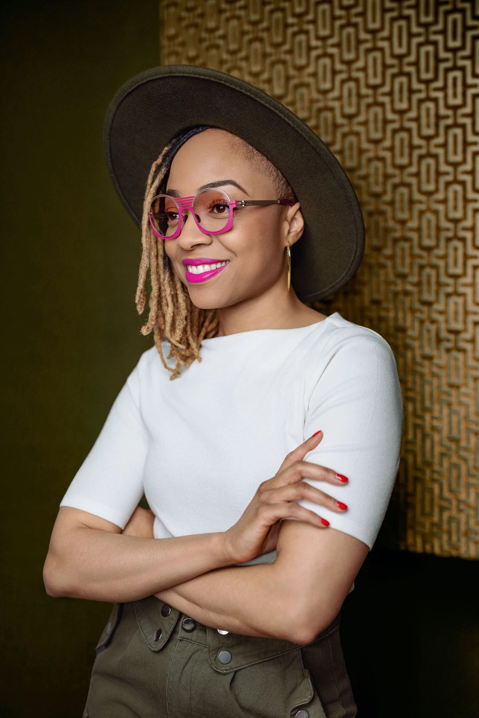 A woman with blonde dreadlocks wearing pink glasses, a black wide-brim hat, a white top, and olive green pants, standing with arms crossed against a patterned background.