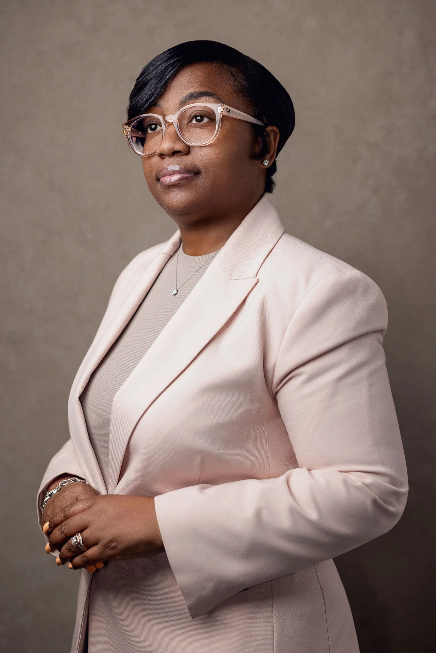 Professional woman with short black hair, wearing glasses and a light pink blazer, standing against a neutral background.