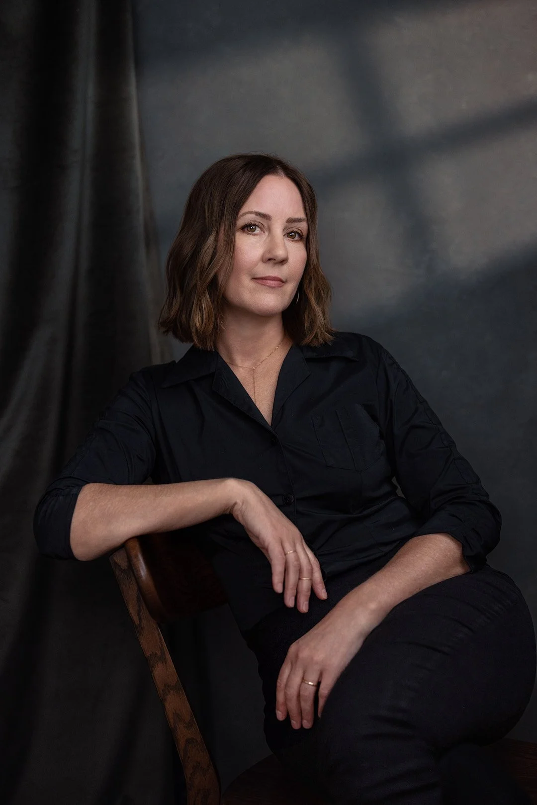 A woman with shoulder-length brown hair, wearing a black shirt, sitting on a wooden chair against a dark backdrop.