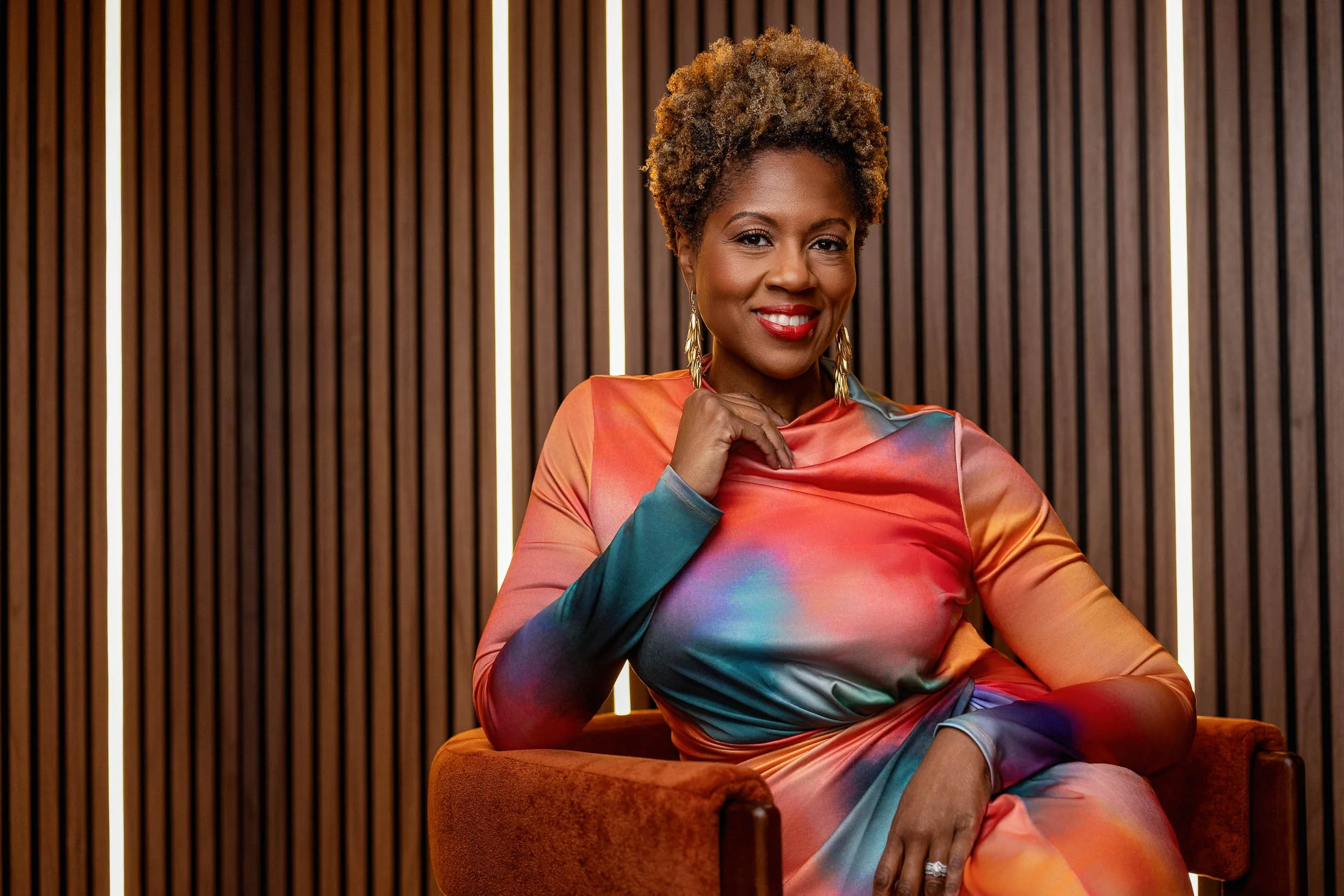 A woman with short curly hair smiling, wearing a colorful, abstract-patterned dress, sitting on an orange chair in front of a wooden slat wall with vertical LED lights.