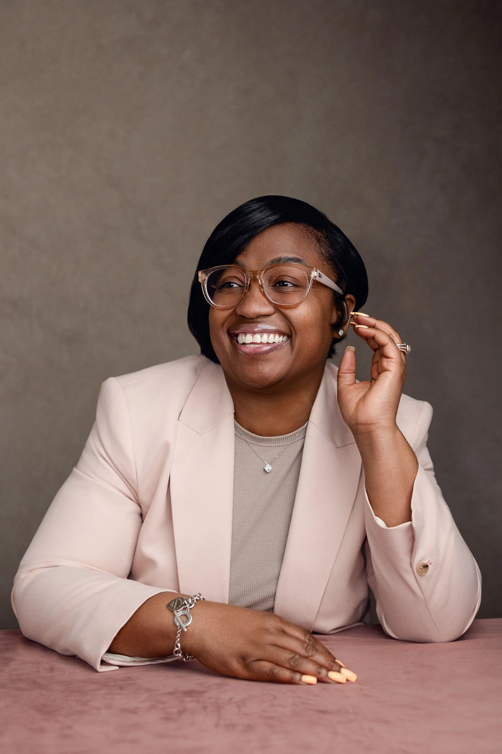 A smiling woman with glasses, wearing a pale pink blazer and jewelry, sitting at a pink table against a gray background.