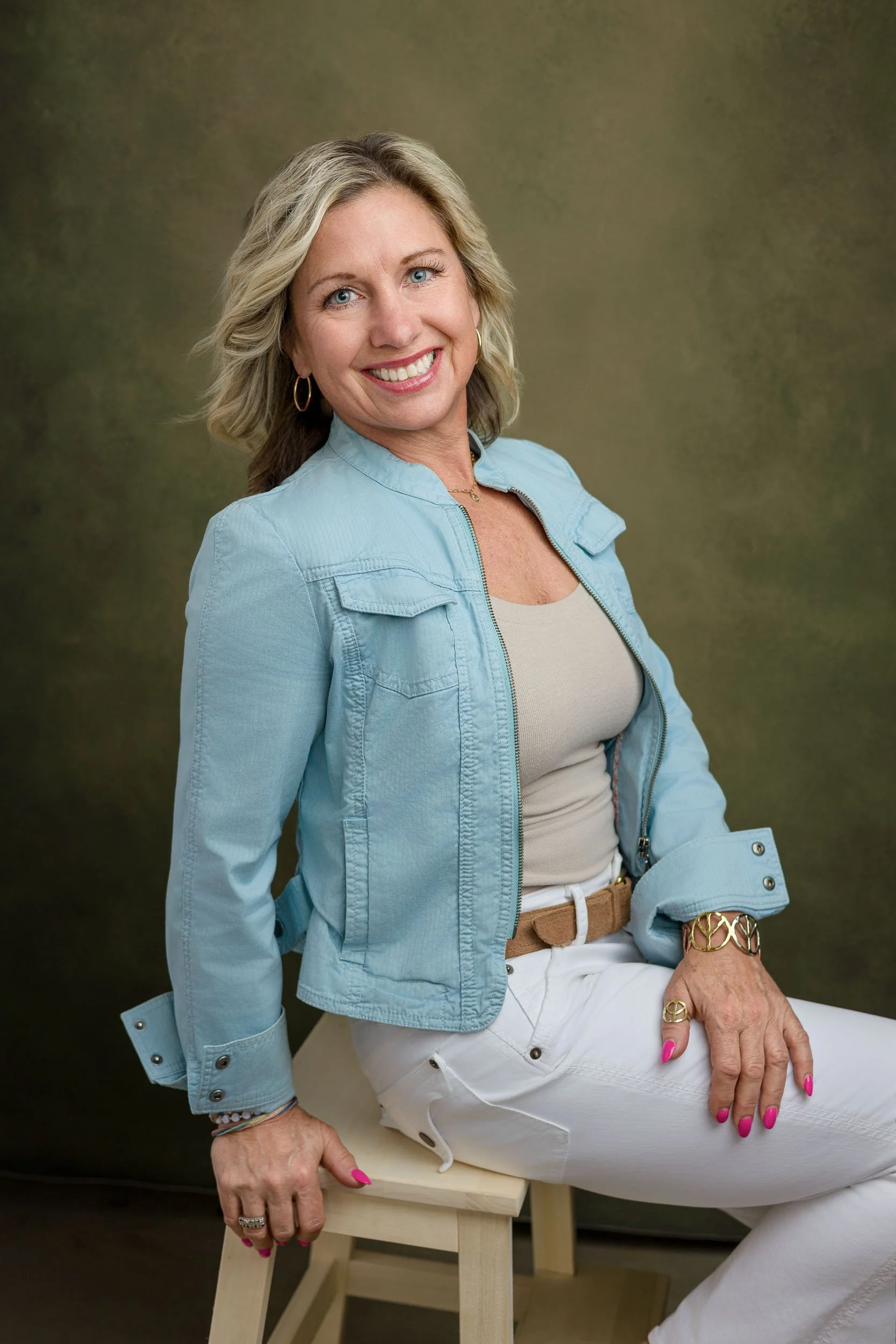 Smiling woman with blonde hair in a casual blue jacket and white pants, sitting on a wooden chair against a dark background.