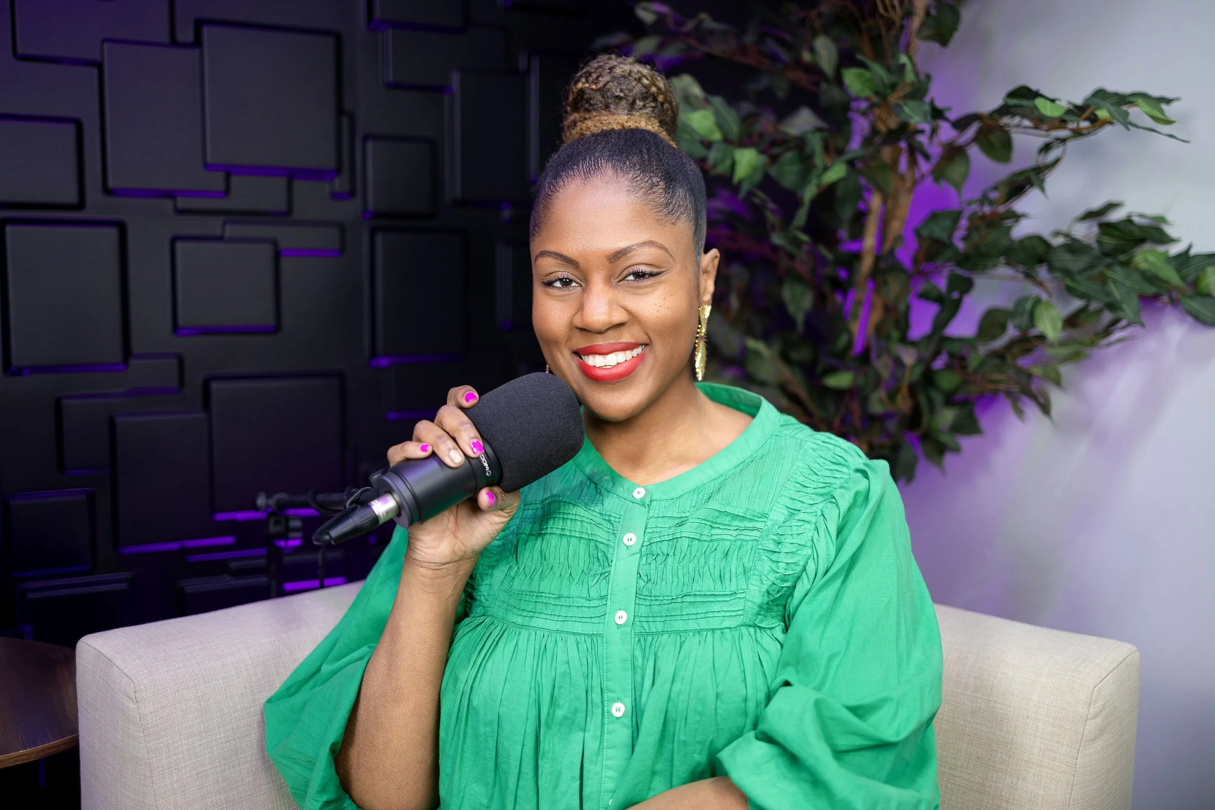A woman with a high bun hairstyle, gold earrings, and red lipstick, holding a microphone and smiling at the camera, sitting on a beige couch in a studio with black acoustic panels and a large green plant in the background.