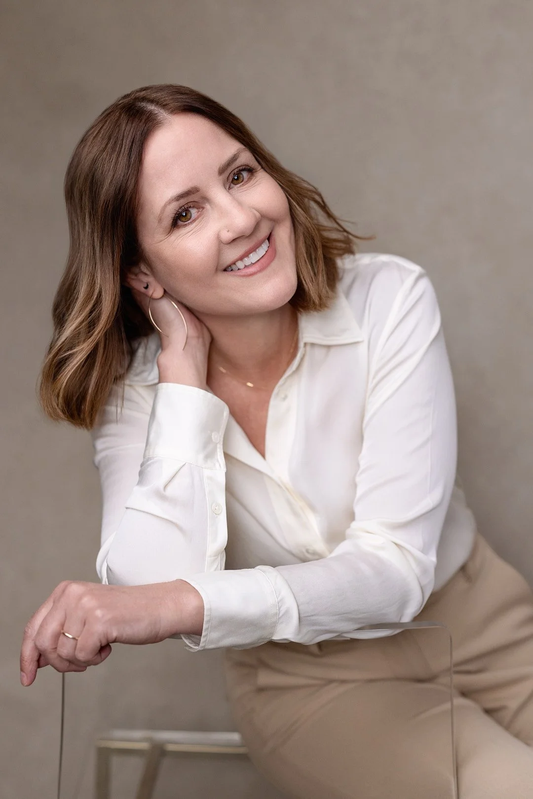 A woman with shoulder-length brown hair, light skin, and wearing a white shirt and beige pants, sitting on a stool against a plain background, smiling at the camera.