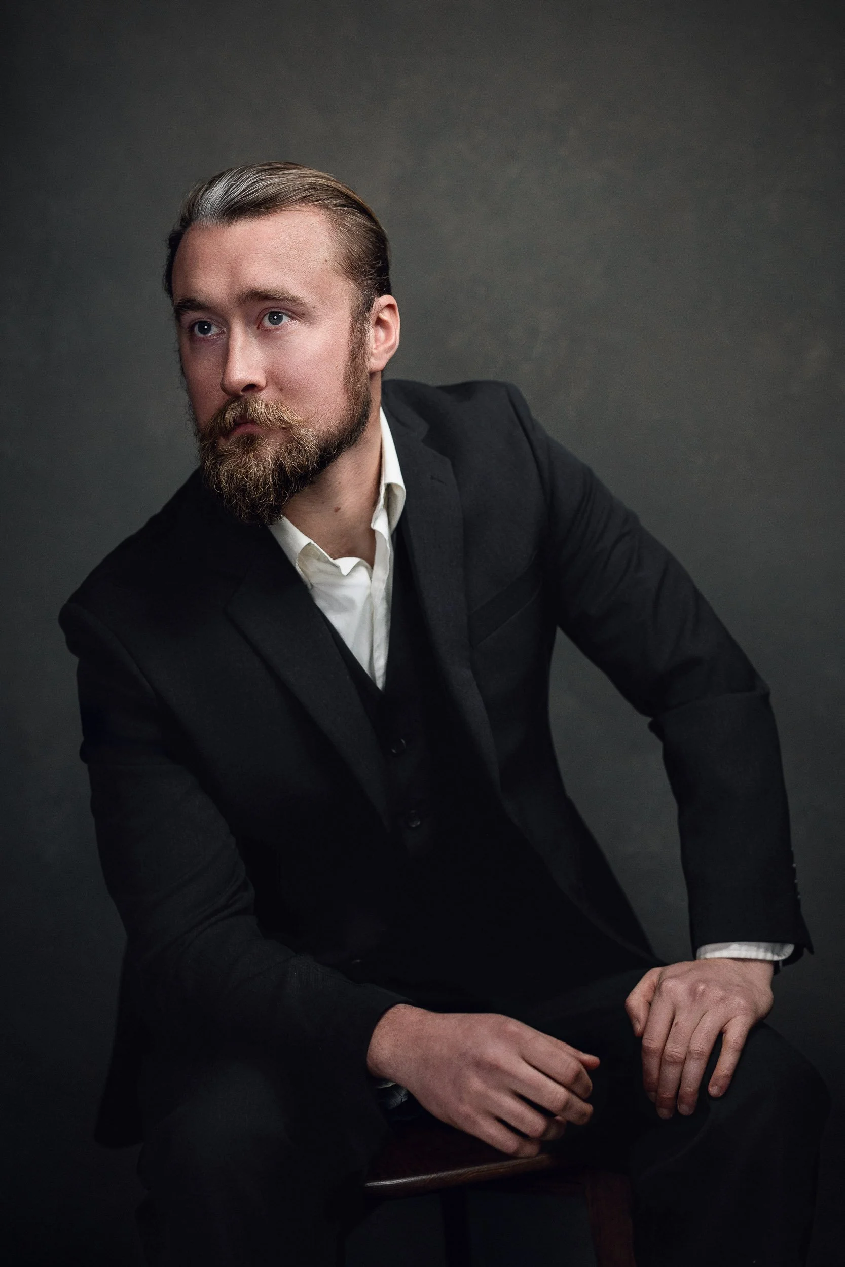 A man with a beard and mustache, wearing a black suit and white shirt, sitting on a stool against a dark background.