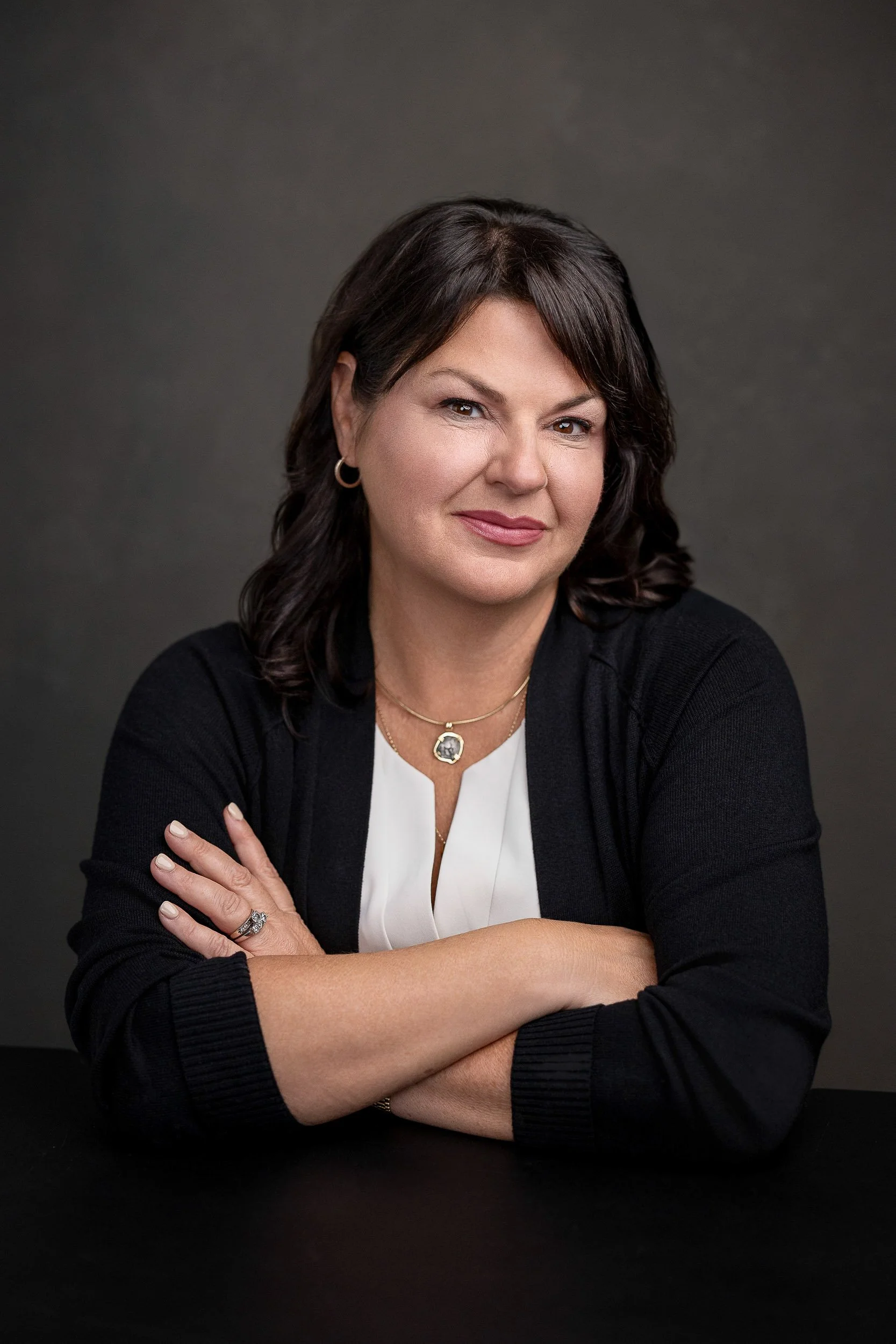 Portrait of a woman with dark brown hair styled in loose waves, wearing a black cardigan over a white top, gold jewelry, and sitting with arms crossed against a dark background.