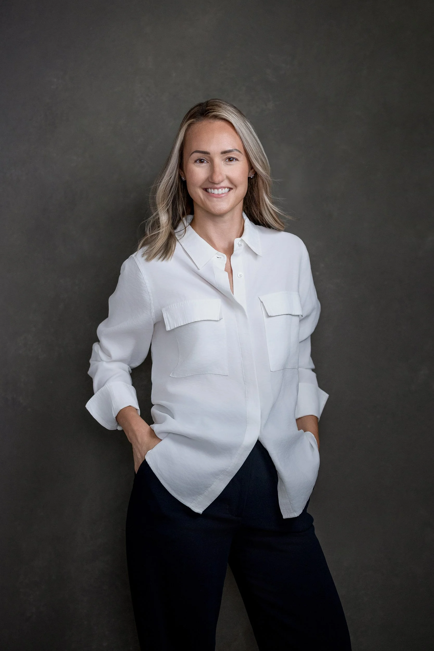 A woman with shoulder-length blonde hair, wearing a white button-up shirt and dark pants, standing against a dark gray background, smiling with hands in pockets.