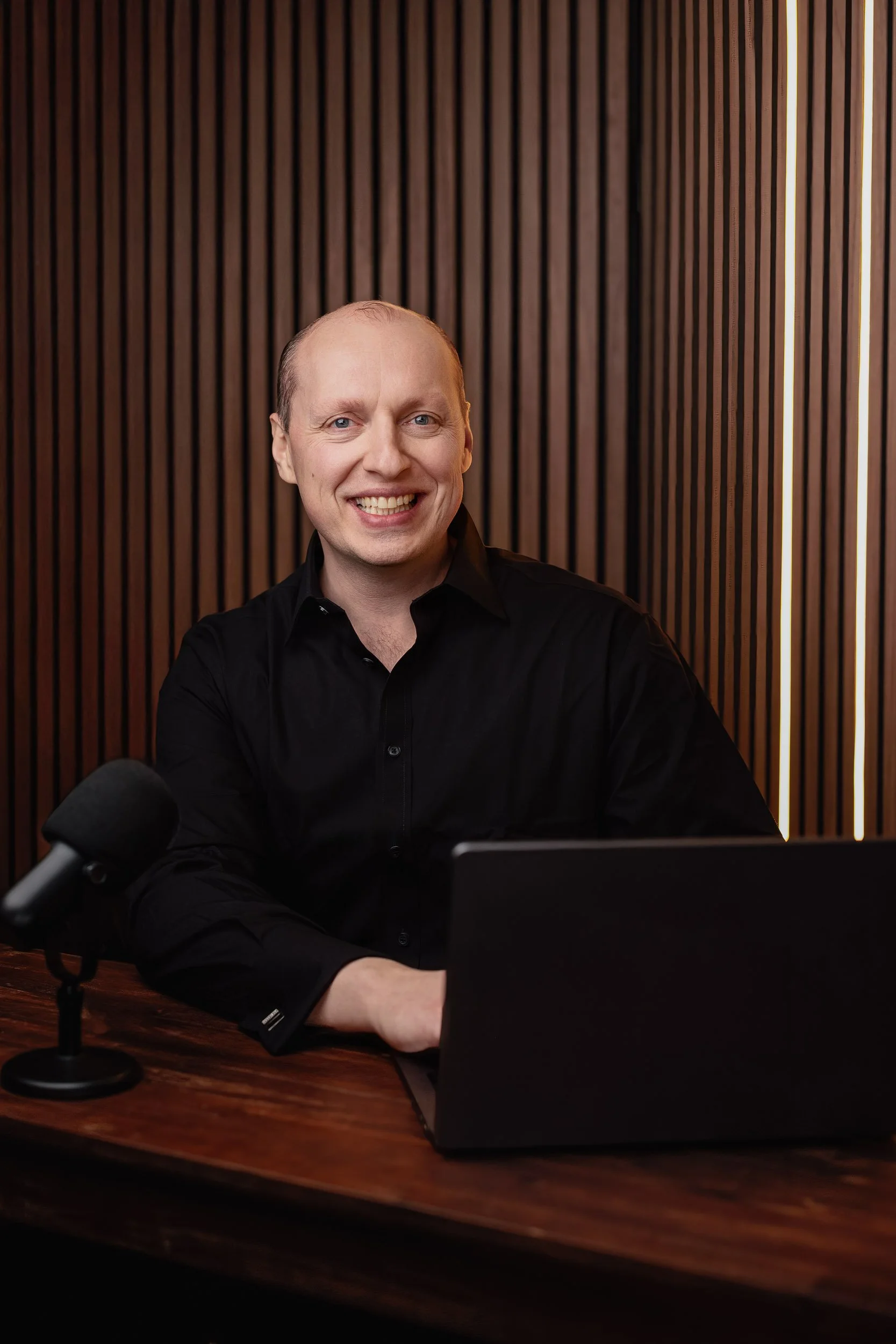 A man with a bald head and blue eyes, smiling, sitting at a wooden table with a laptop and a microphone, against a wood-paneled wall.