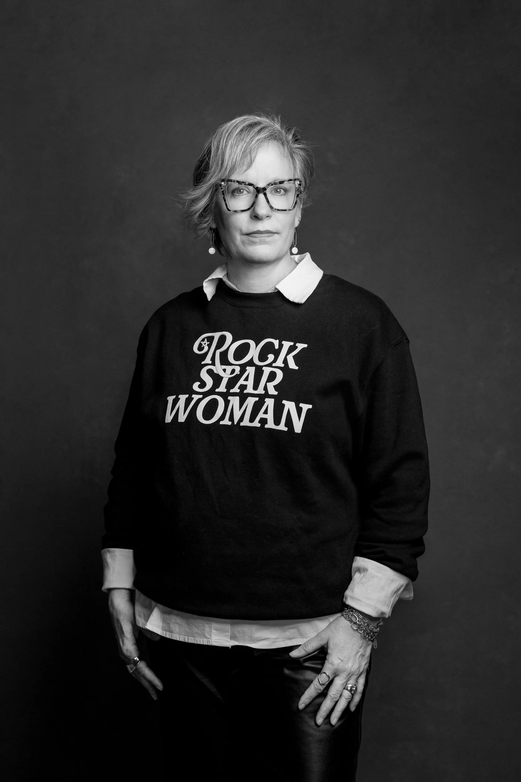 A woman wearing glasses and earrings, dressed in a layered outfit with a sweatshirt that says "Rock Star Woman," standing against a plain background.