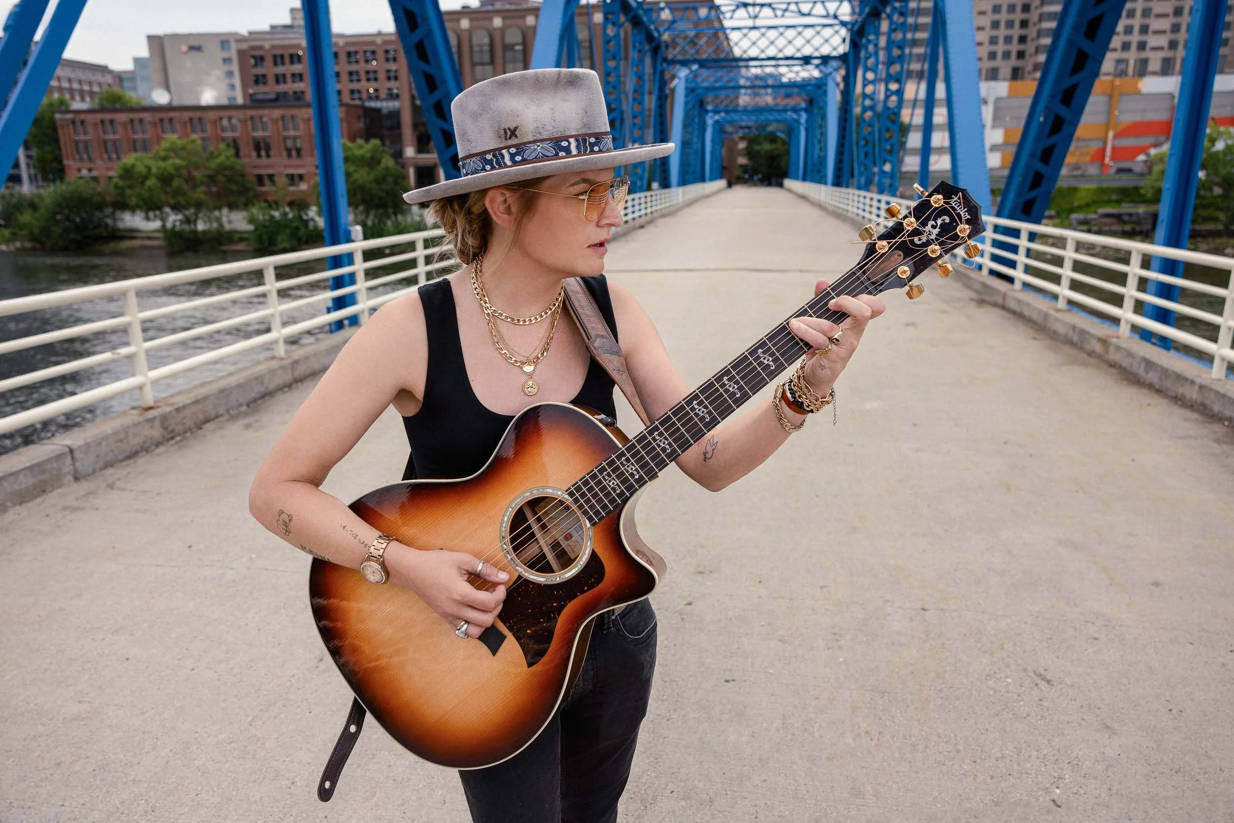 A woman with blonde hair, wearing a gray hat, sunglasses, black tank top, and gold jewelry, stands on an outdoor bridge while playing an acoustic guitar.