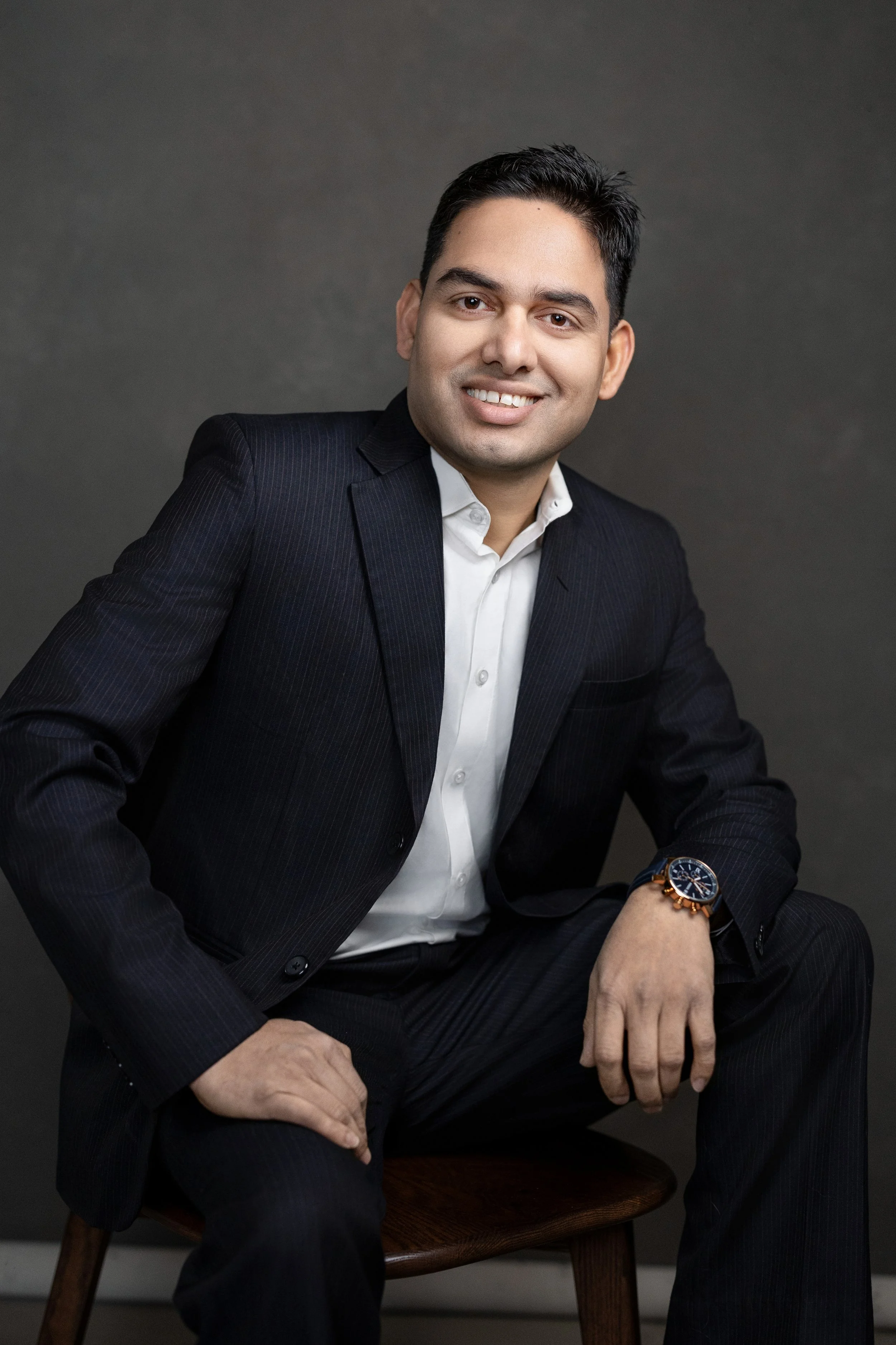 A man in a dark suit and white shirt sitting on a wooden chair, smiling at the camera with a gray background.