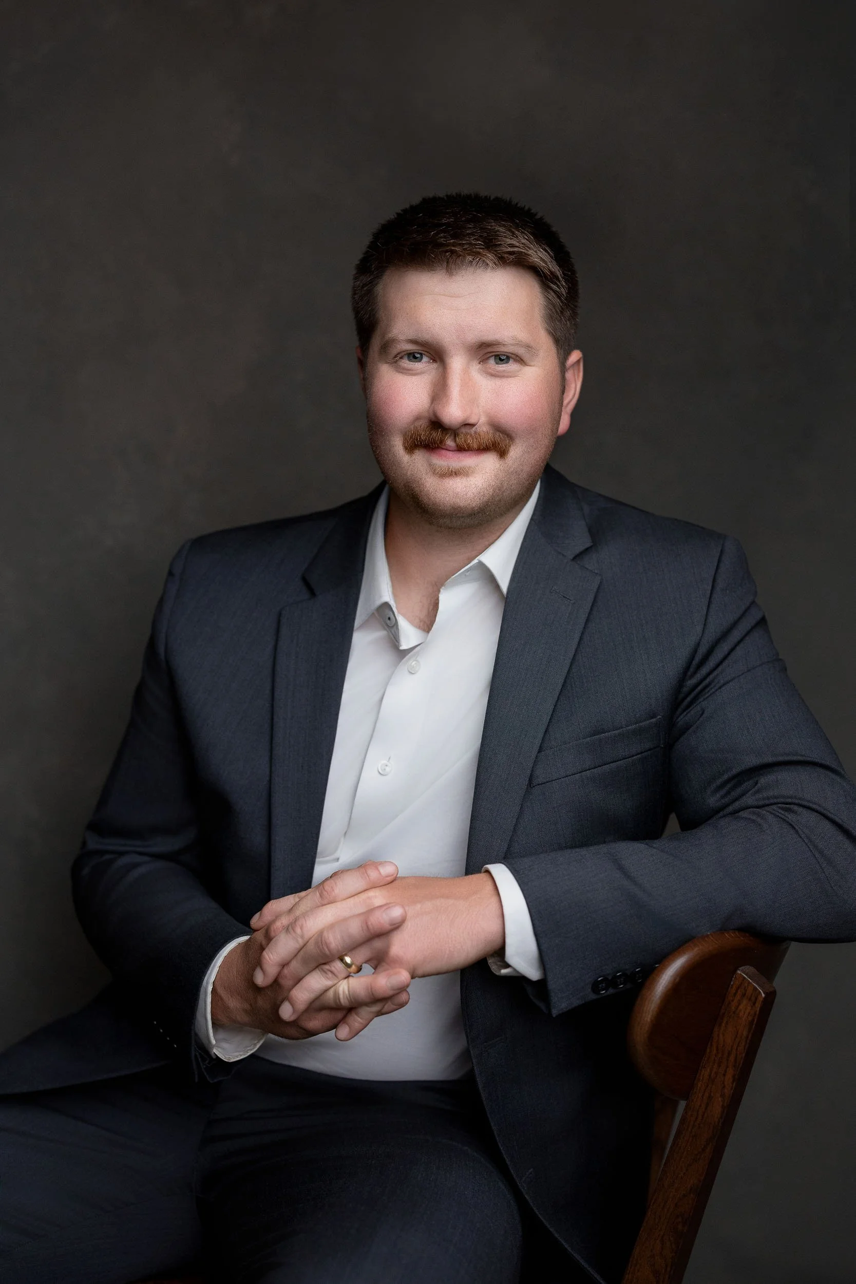 A man with brown hair, a mustache, and blue eyes, wearing a dark suit and white shirt, sitting on a wooden chair against a dark background.