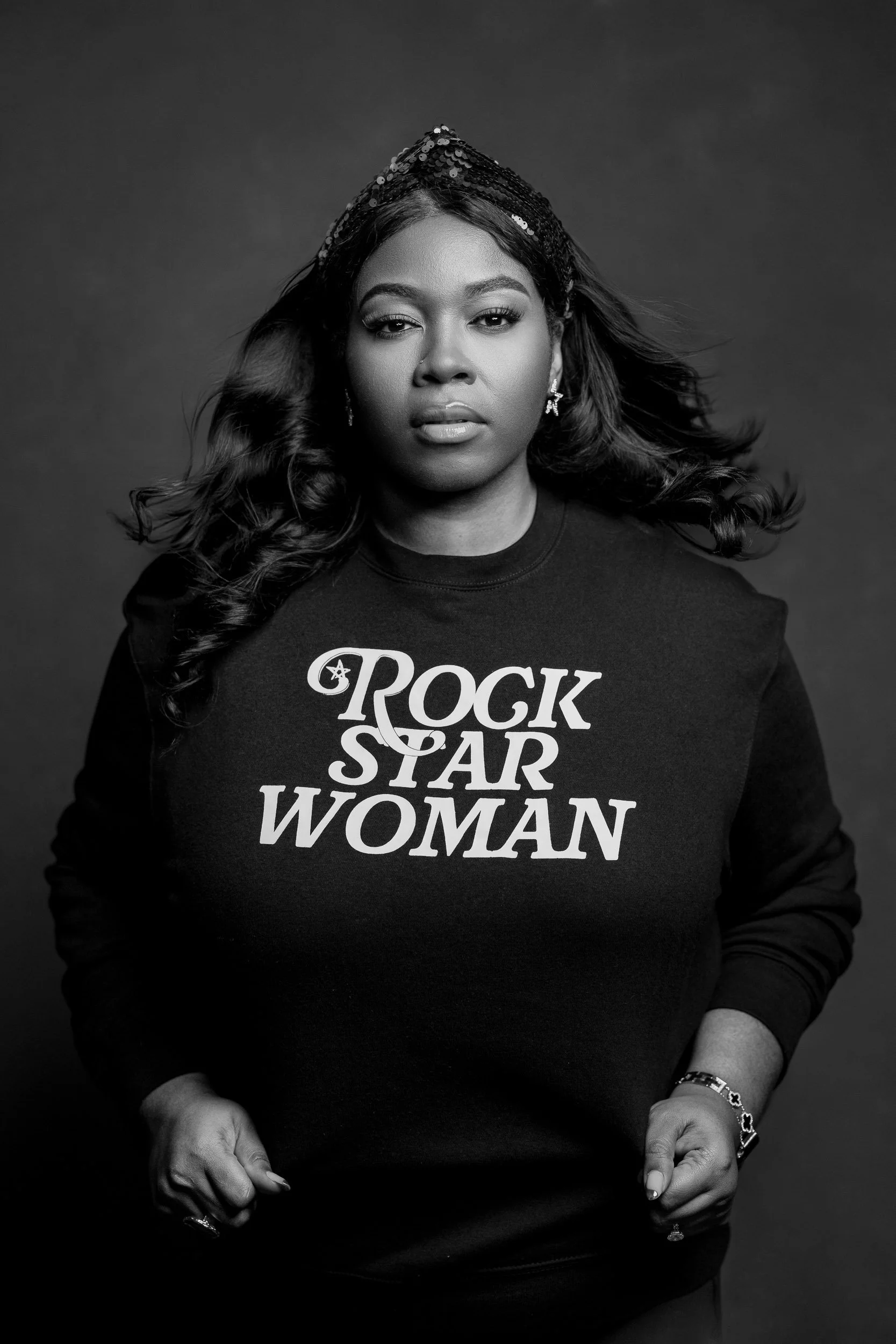 Black and white portrait of a woman wearing a sweatshirt that says 'ROCK STAR WOMAN' and a shiny headband, looking directly at the camera with her hands in her pockets.