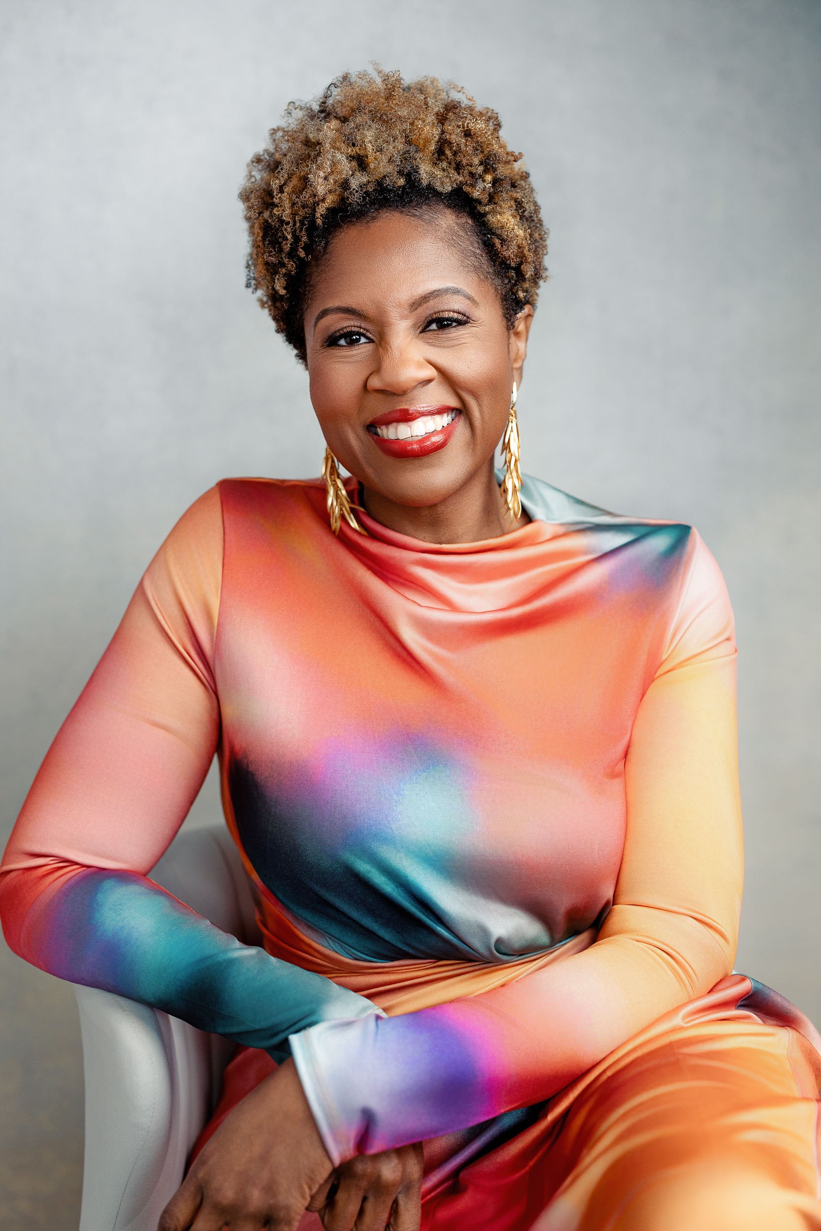A woman with curly short hair smiling, wearing a colorful, tie-dye dress and gold earrings, seated against a neutral background.