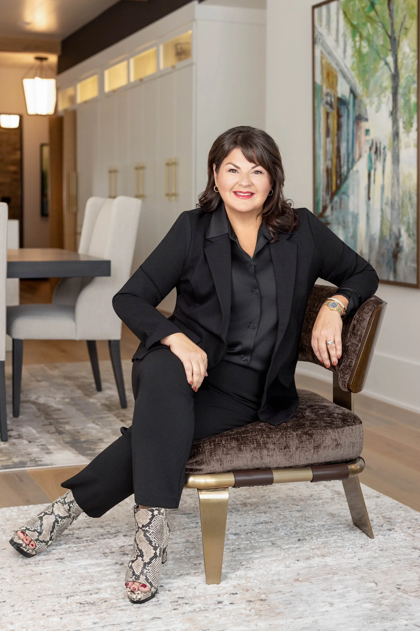 A woman in black clothing and snakeskin boots seated on a chair in a modern, well-lit room with a colorful painting on the wall behind her.