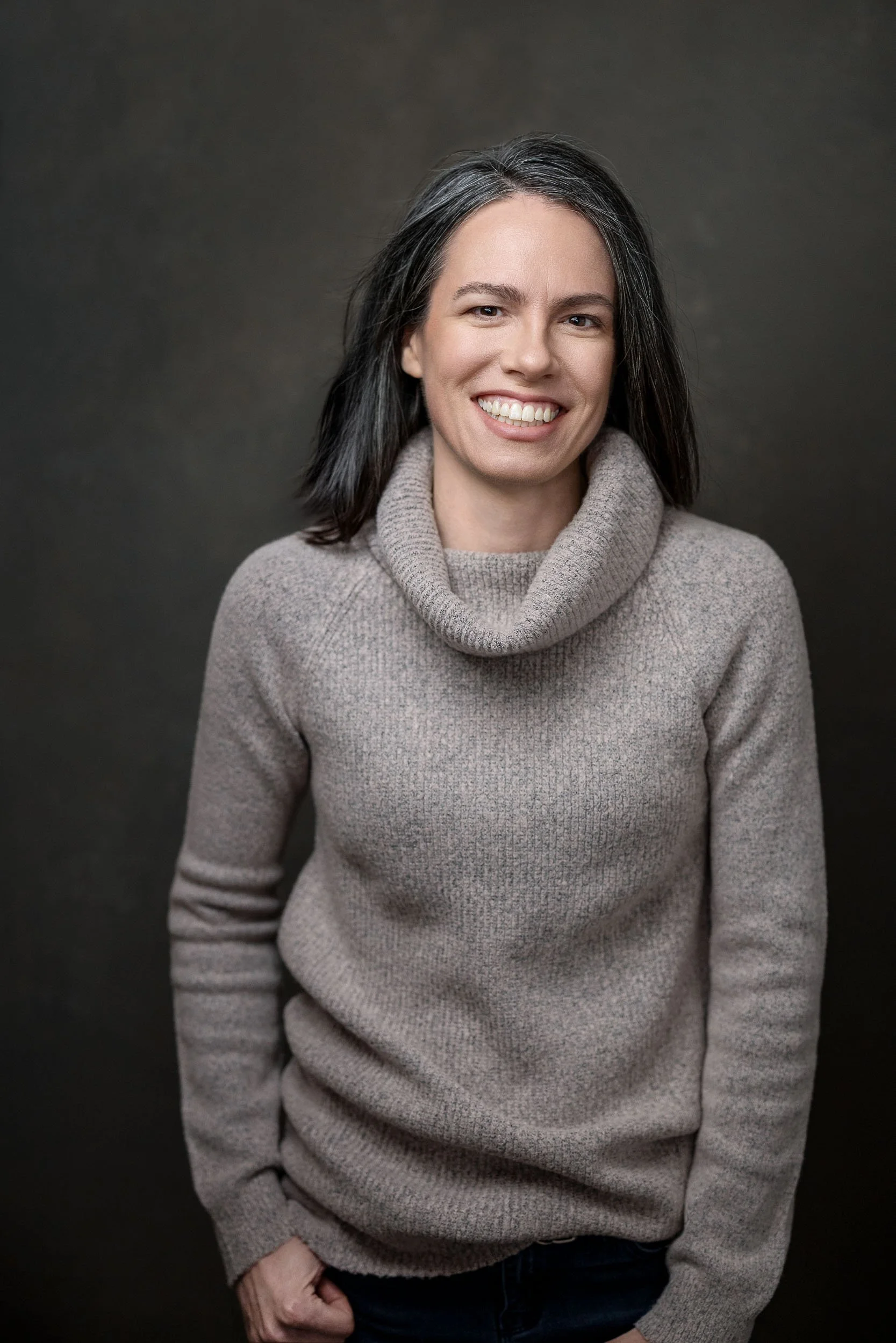 A woman with dark hair smiling, wearing a gray turtleneck sweater, against a dark background.