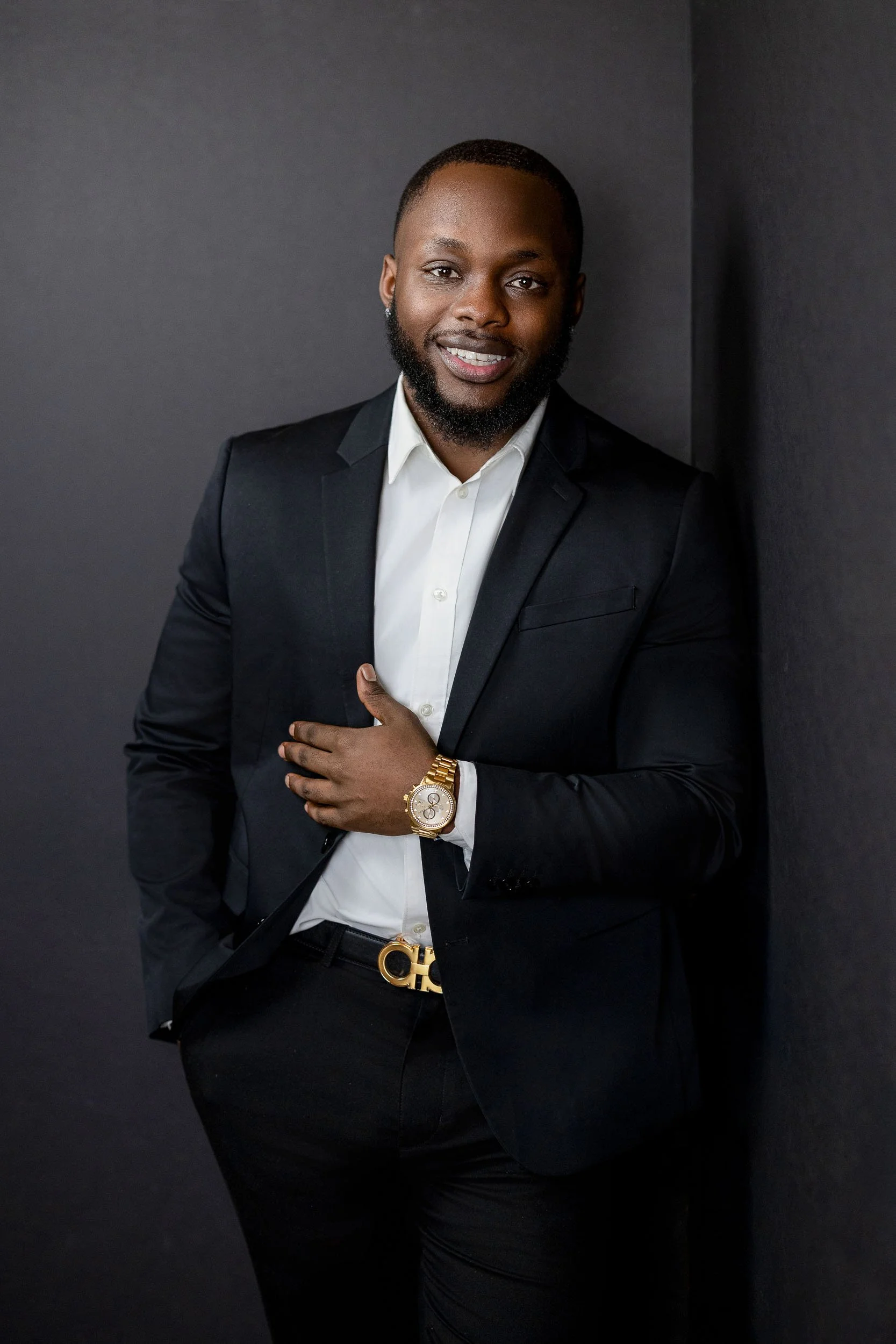 A man in a black suit and white shirt, smiling, standing against a dark gray wall, wearing a gold watch and belt.