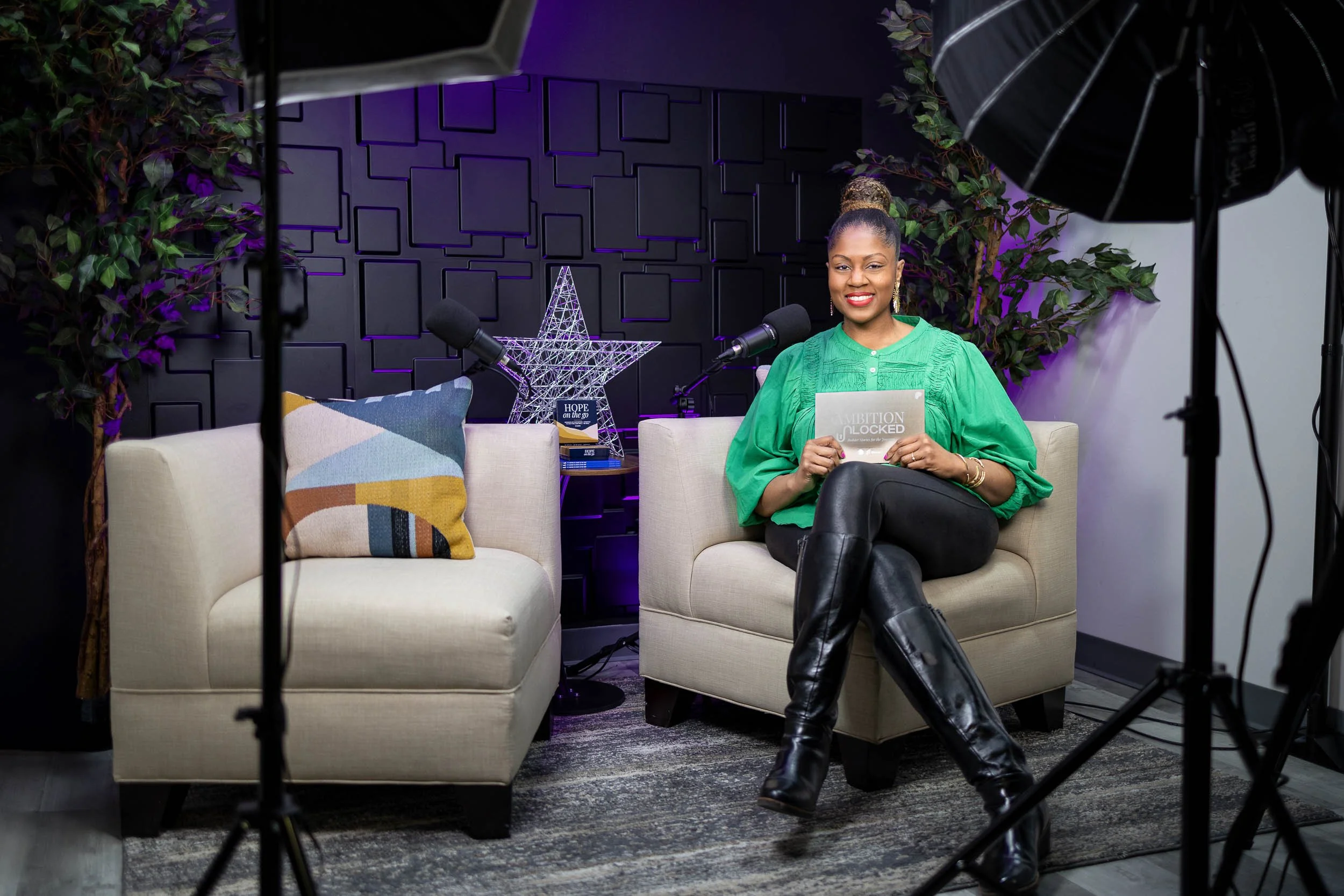 A woman sitting on a beige couch holding a book, surrounded by photography equipment in a studio setting with decorative lights and plants.