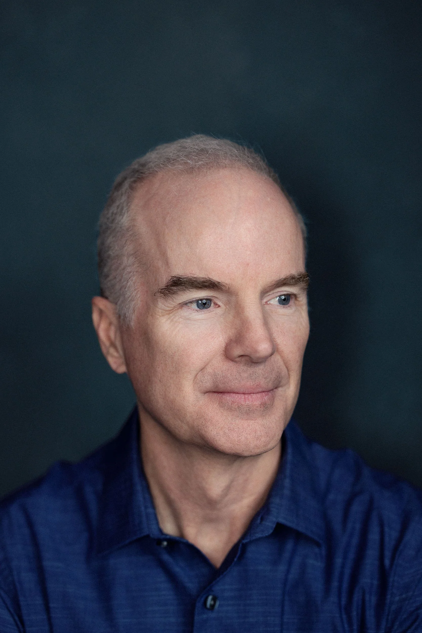 Close-up portrait of a middle-aged man with short gray hair, blue eyes, and fair skin, wearing a dark blue collared shirt, against a dark backdrop.