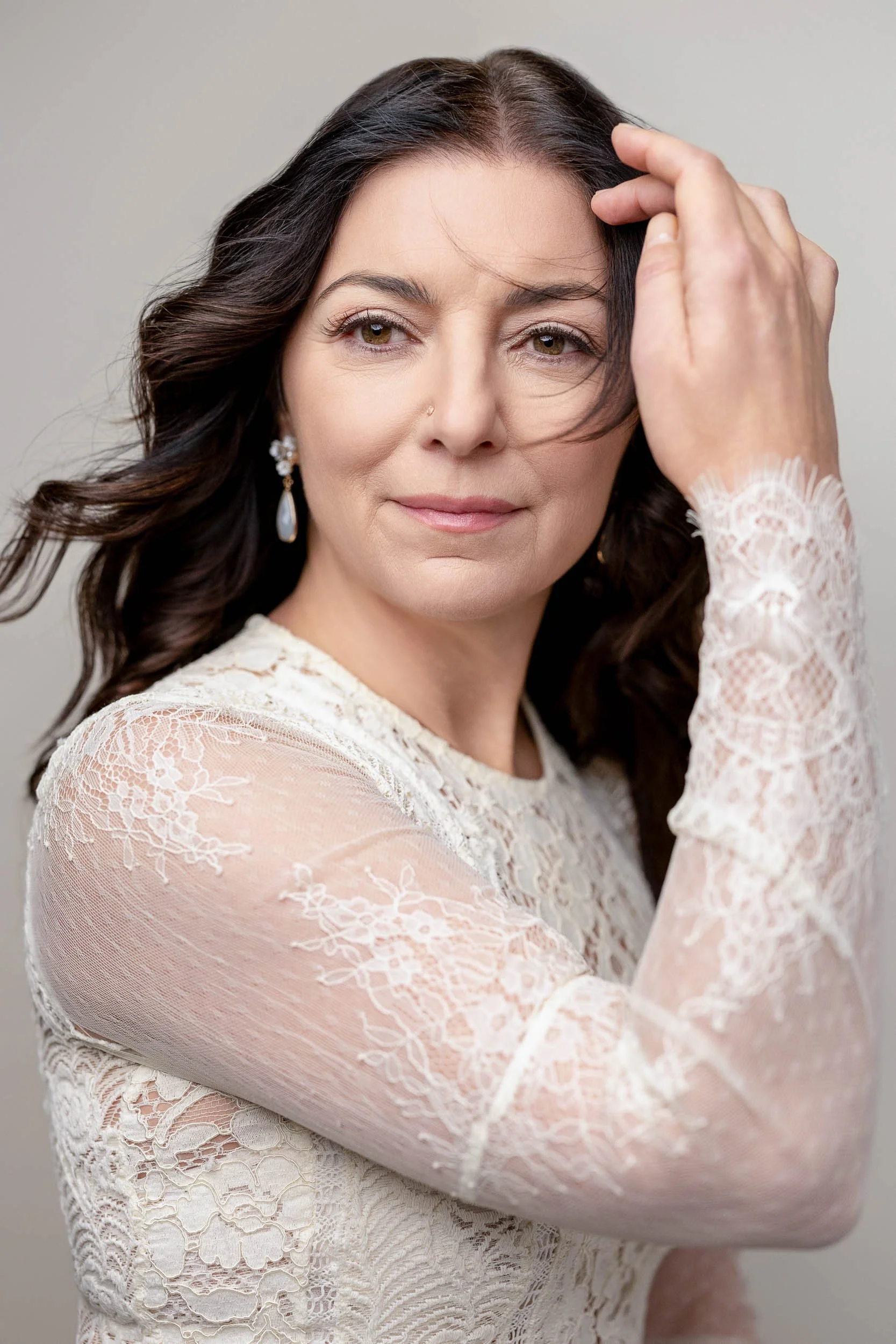 A woman with long dark hair, wearing a white lace dress and earrings, is touching her forehead and looking softly at the camera against a plain backdrop.