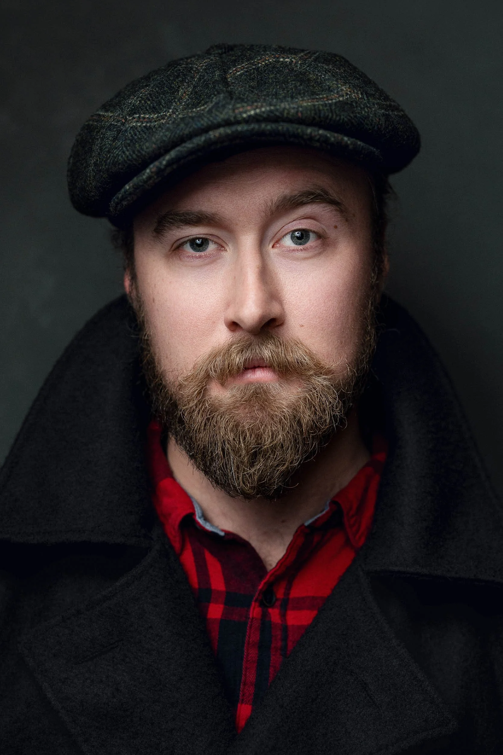 A portrait of a man with a beard, wearing a plaid flat cap, red and black flannel shirt, and dark coat, against a dark background.