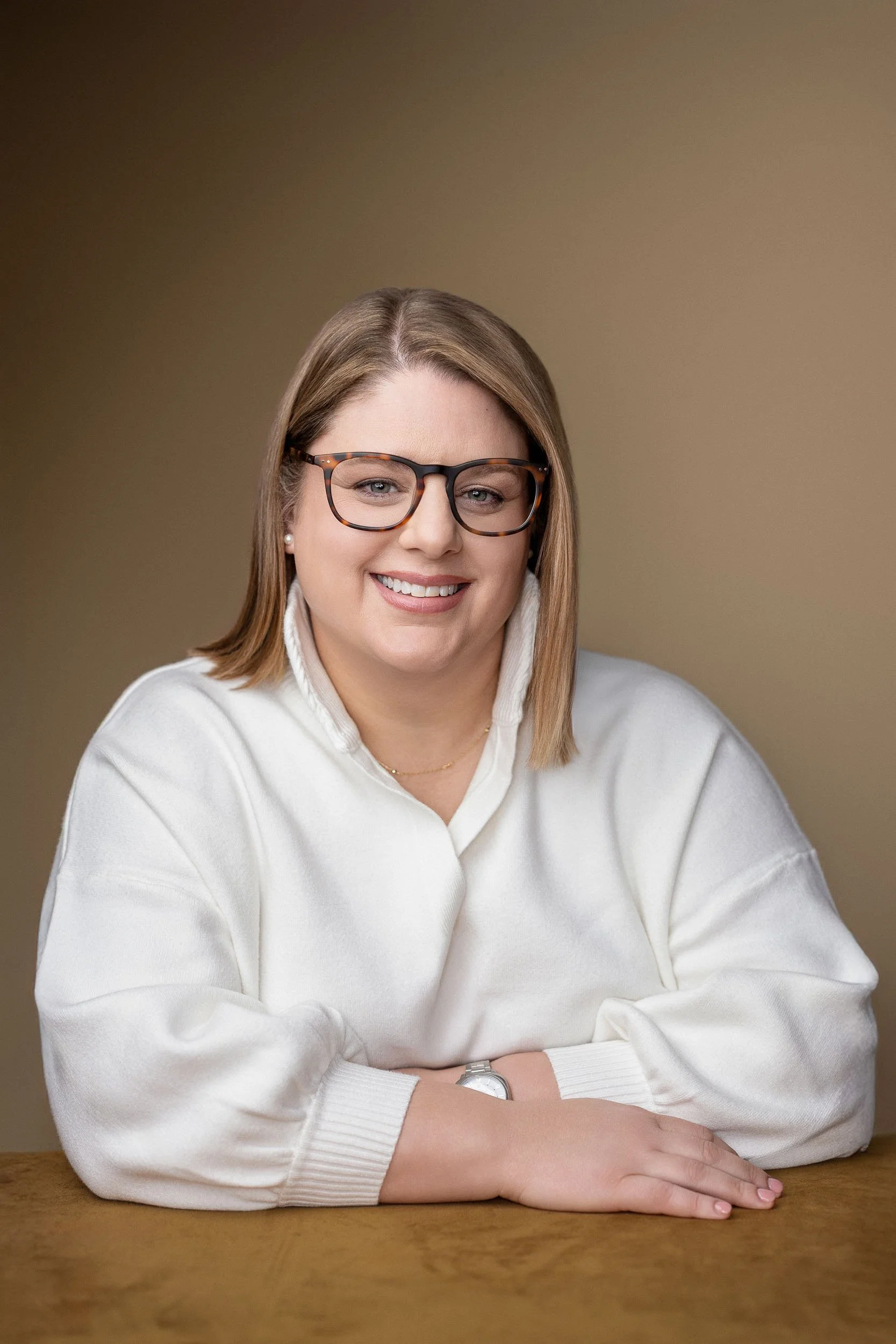 A woman with shoulder-length blonde hair, glasses, and a white sweater, smiling and sitting at a wooden table against a beige background.