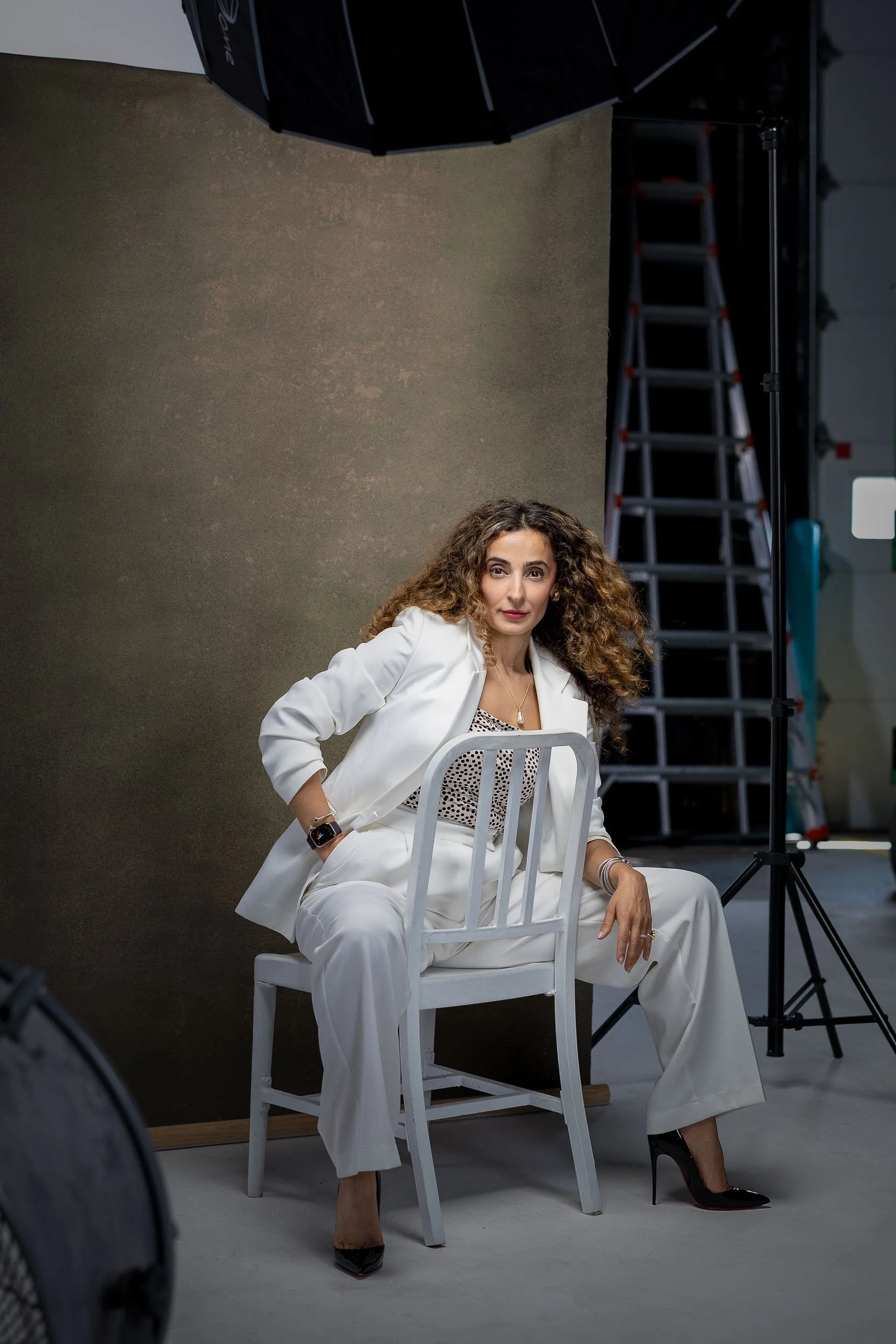 A woman with curly hair and a white suit sitting on a white chair in a photography studio, with lighting and ladder equipment visible in the background.
