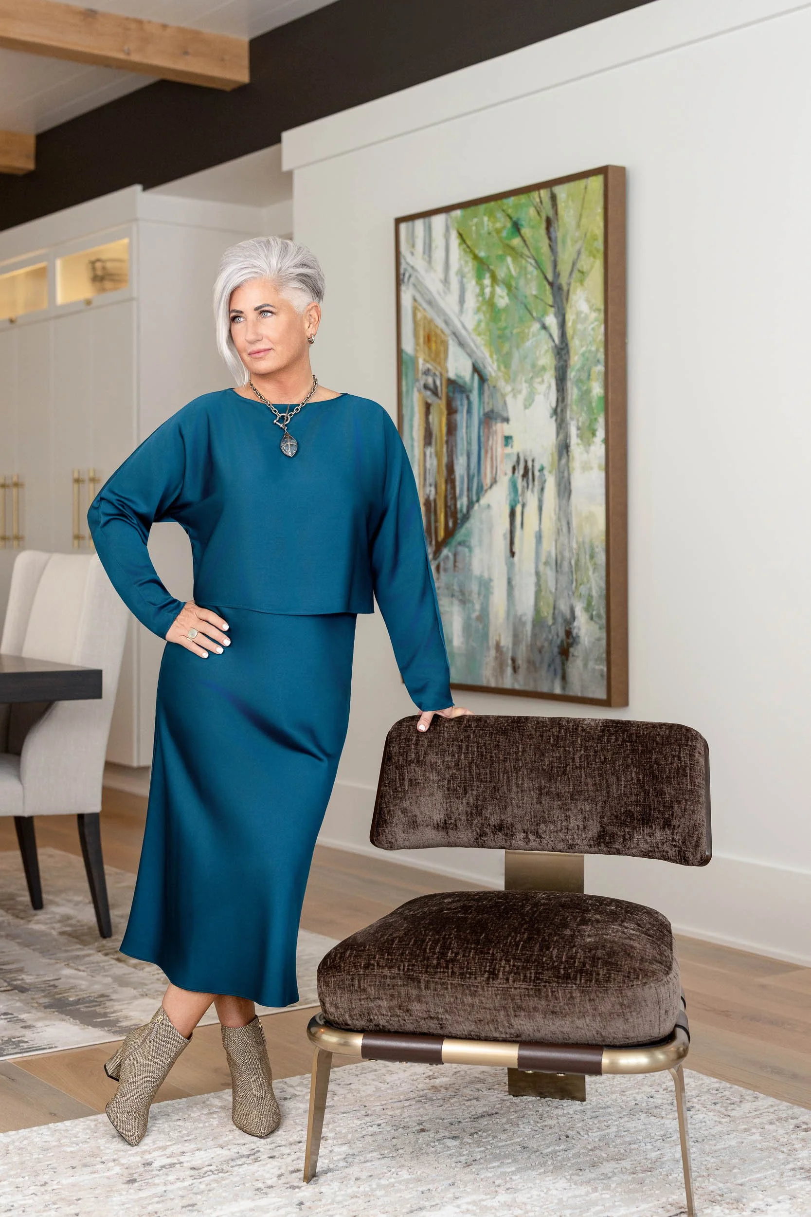 A woman with gray hair wearing a teal dress and ankle boots standing beside a brown upholstered chair in a room with modern decor and a large colorful painting of a street scene on the wall.