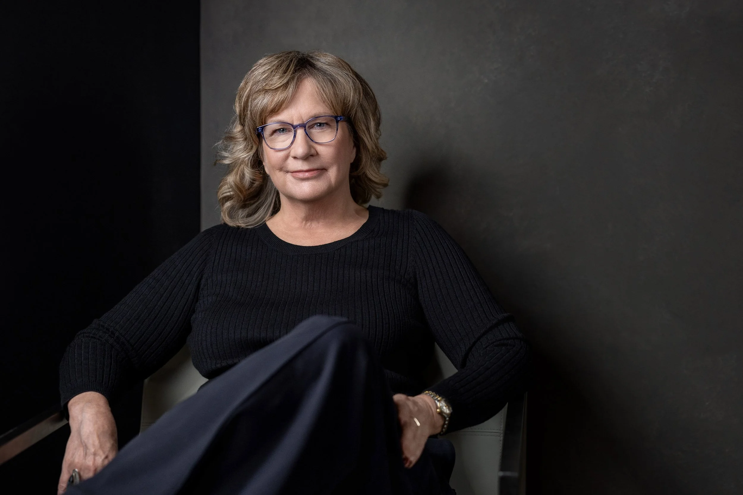 A woman in a long sleeved black shirt sitting on a white chair with metal frame against a dark textured background.