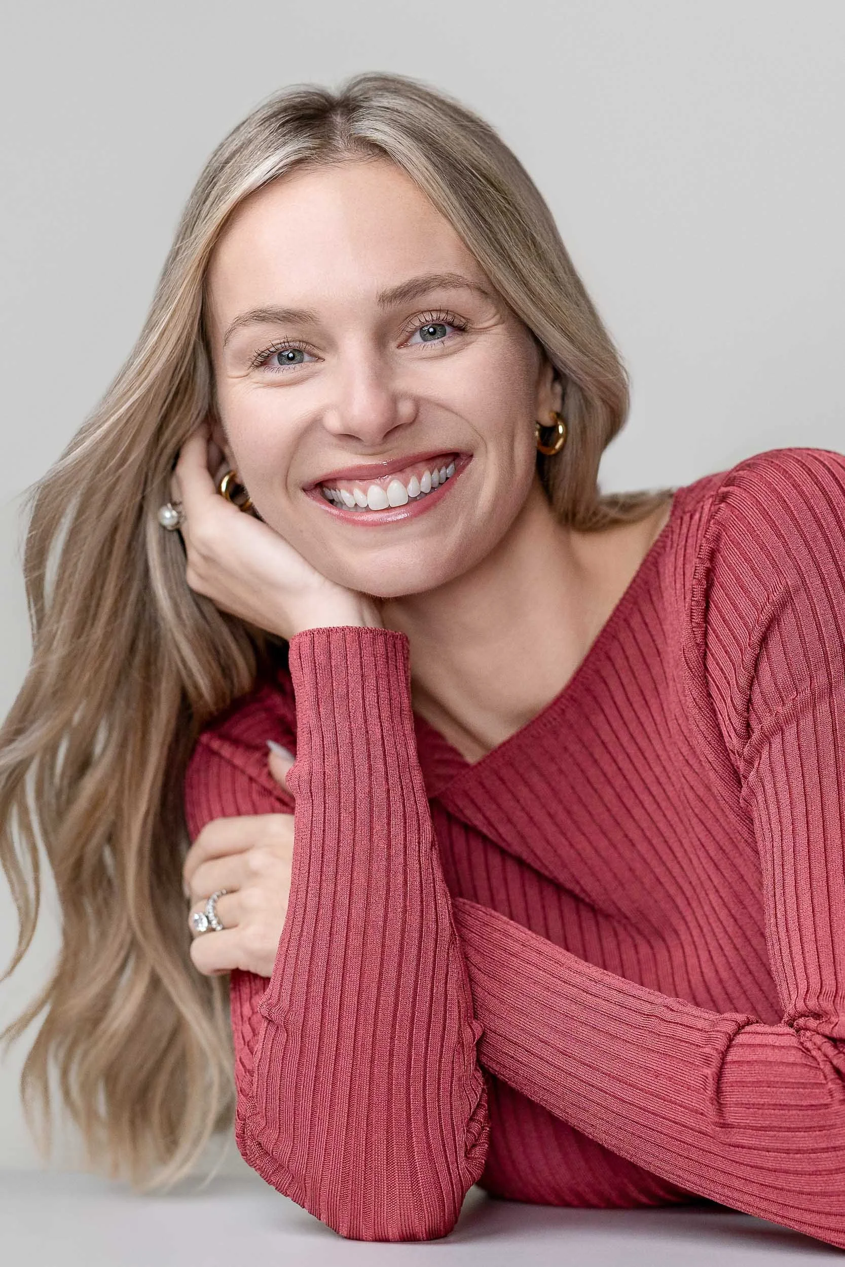 A woman with long blonde hair smiling, wearing a red ribbed long-sleeve top and gold hoop earrings, resting her head on her hand.