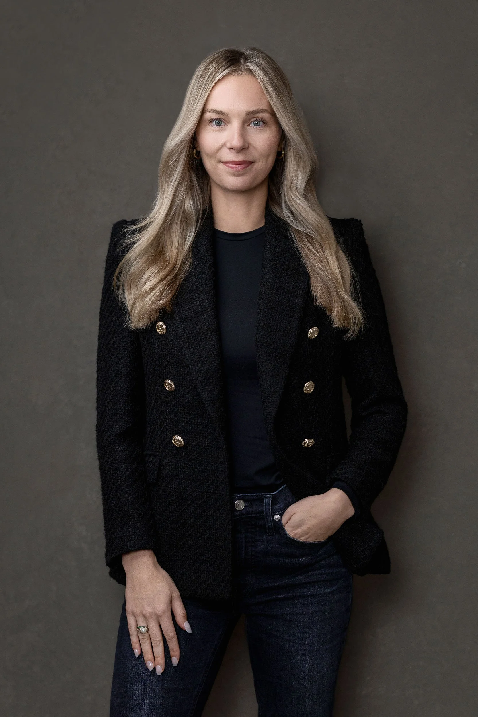 A woman with long blonde hair, wearing a black blazer with gold buttons, black shirt, and dark jeans, standing against a gray background.