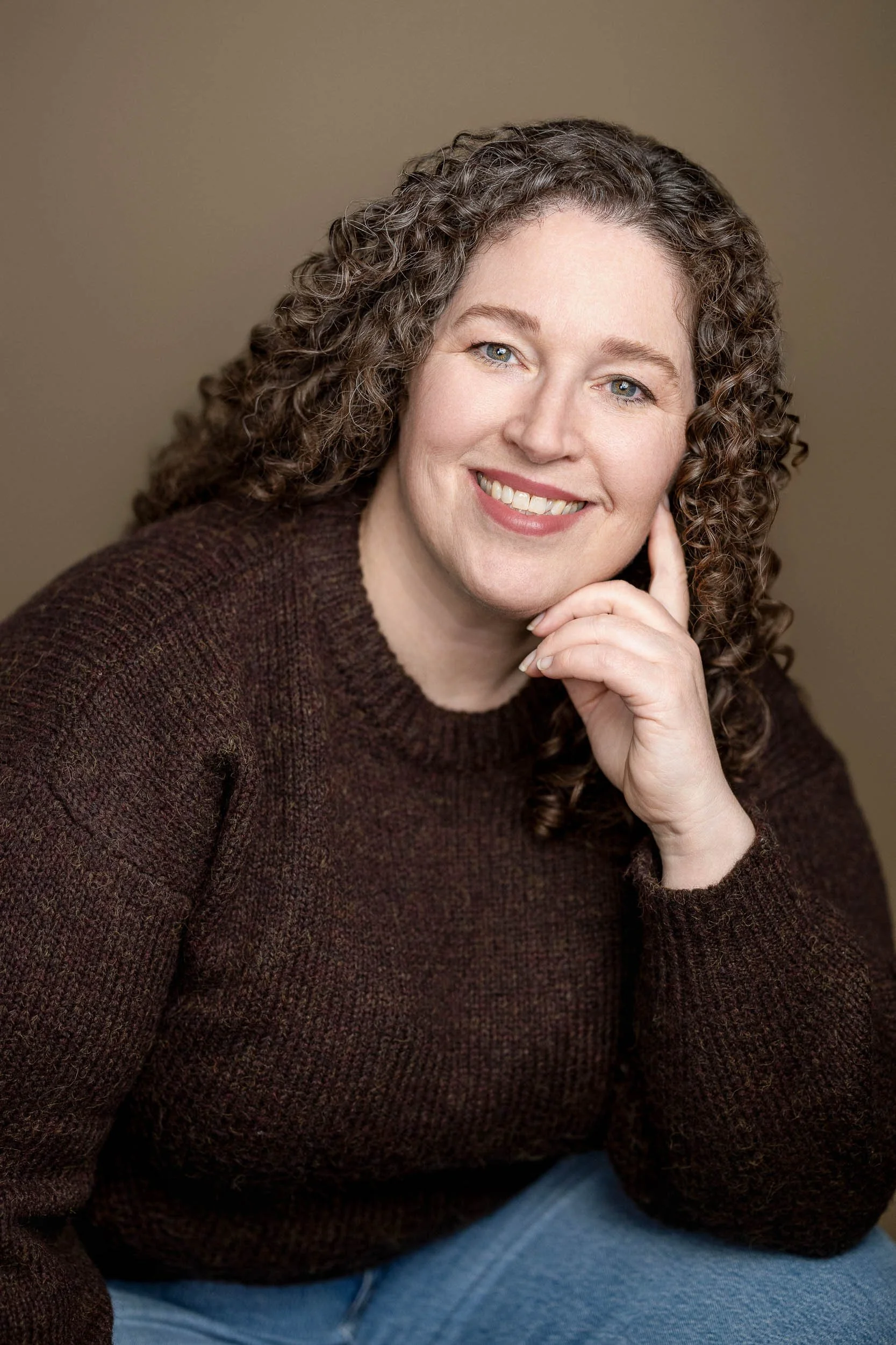 A woman with curly hair wearing a brown sweater, sitting with her hand resting on her face, smiling at the camera against a beige background.