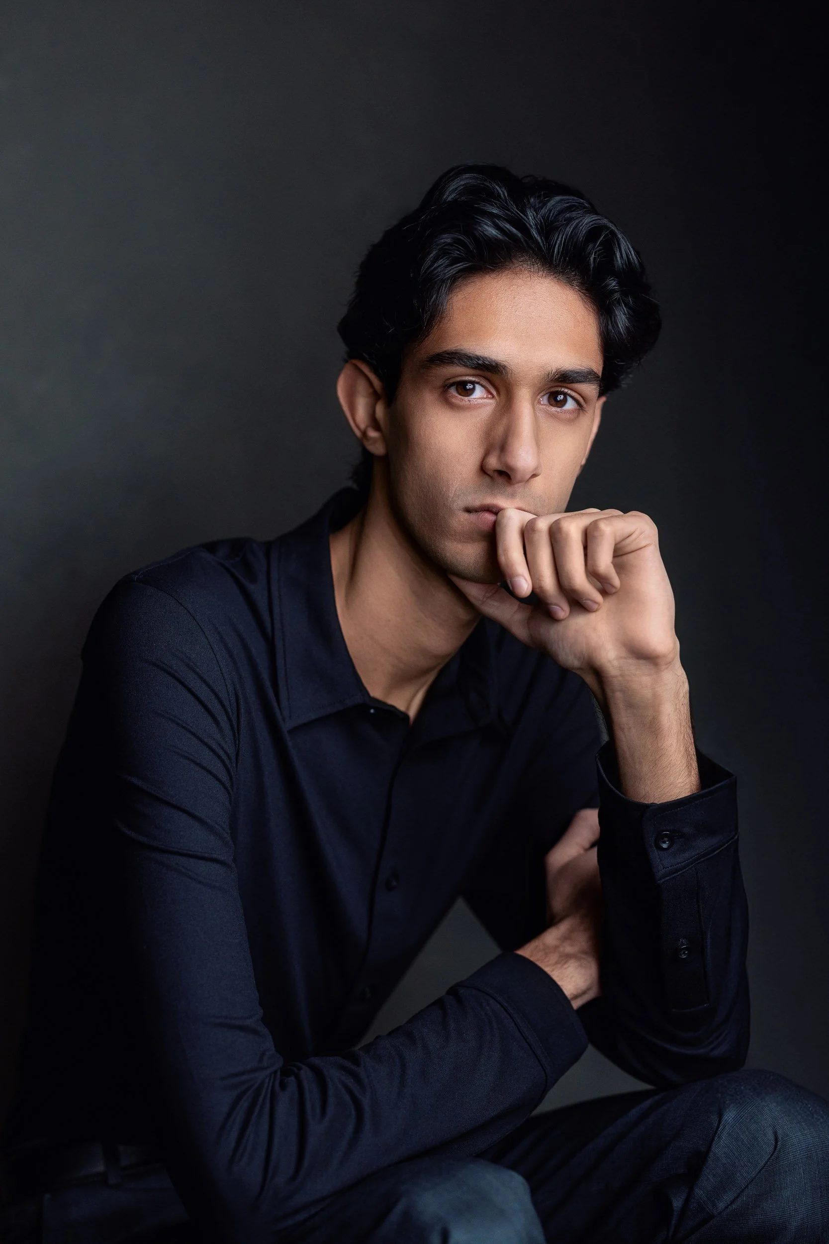 A young man with black hair wearing a black shirt, sitting with his hand near his mouth, looking at the camera against a dark background.