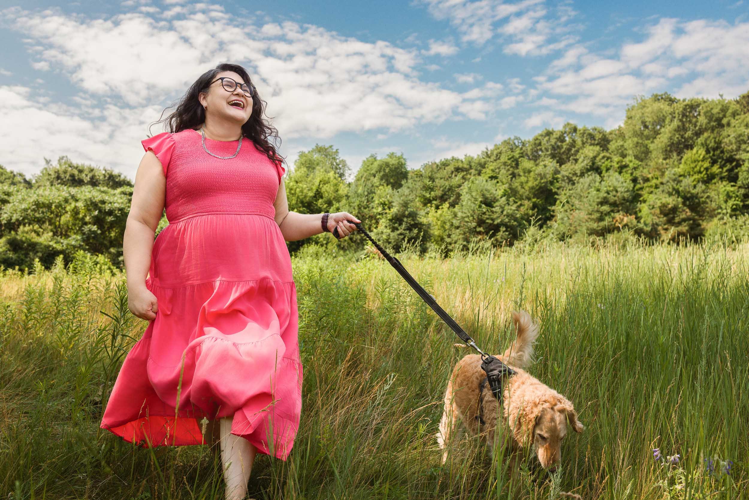 A woman in a pink dress walking a golden retriever dog in a grassy field with trees and a partly cloudy sky in the background.