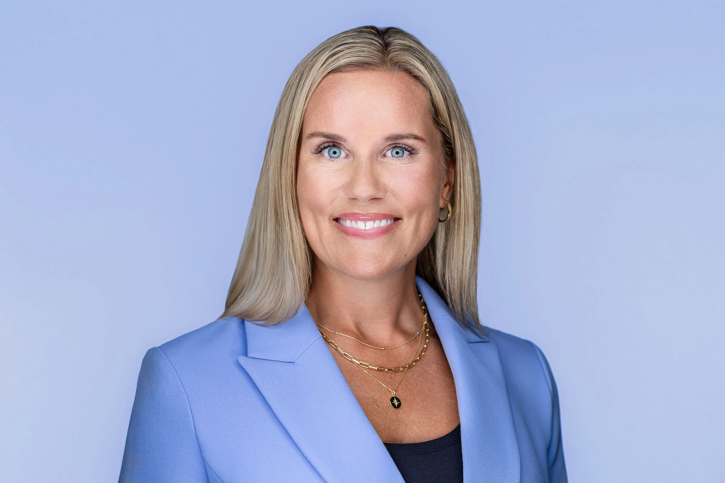 Headshot of a smiling woman with blonde hair wearing a blue blazer and layered gold necklaces against a light blue background.