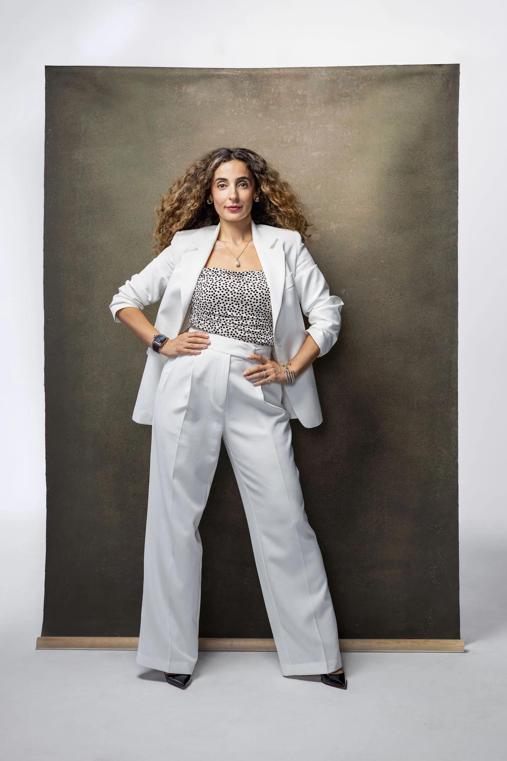 A woman with curly hair wearing a white suit and polka dot top standing with hands on hips in front of a dark backdrop.