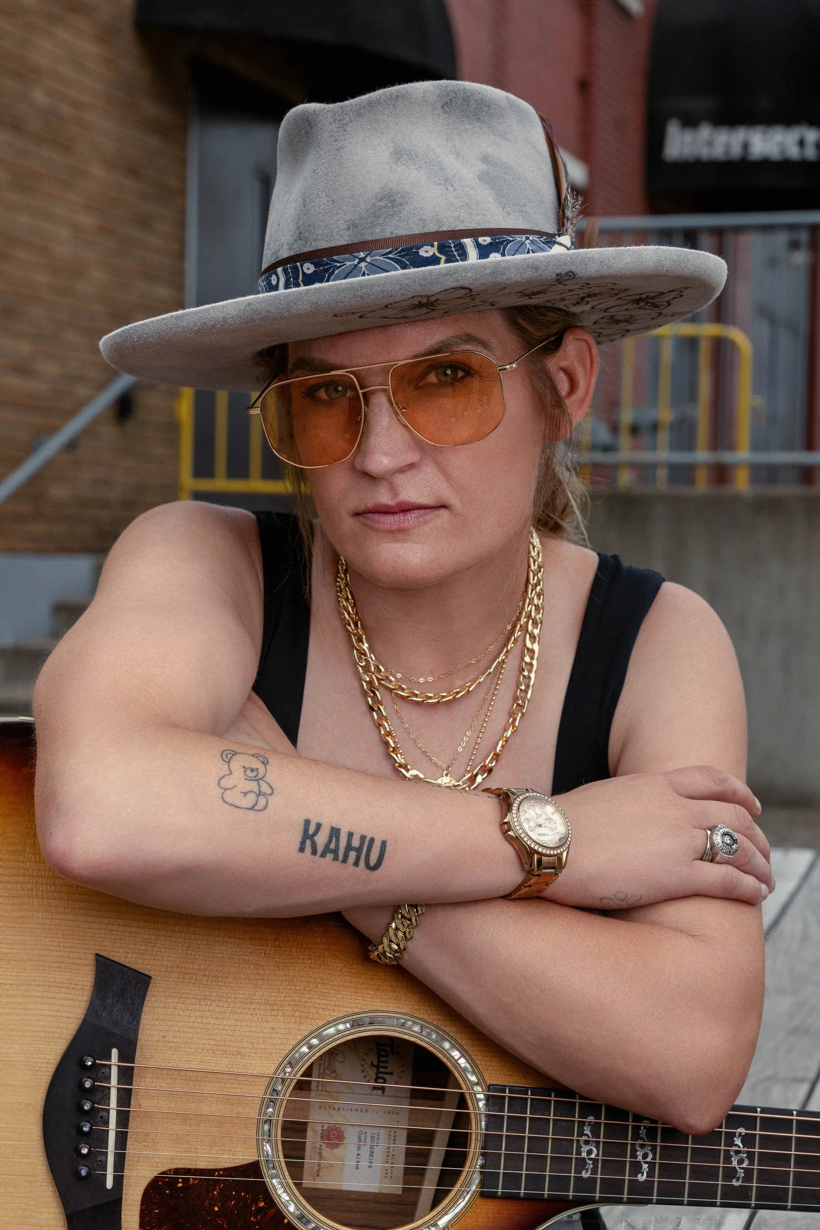 A woman wearing a wide-brimmed hat with a decorative band, orange-tinted sunglasses, multiple gold chains, a wristwatch, a ring, and a bracelet, sitting with her arms crossed on a guitar, outdoors in front of a brick building.