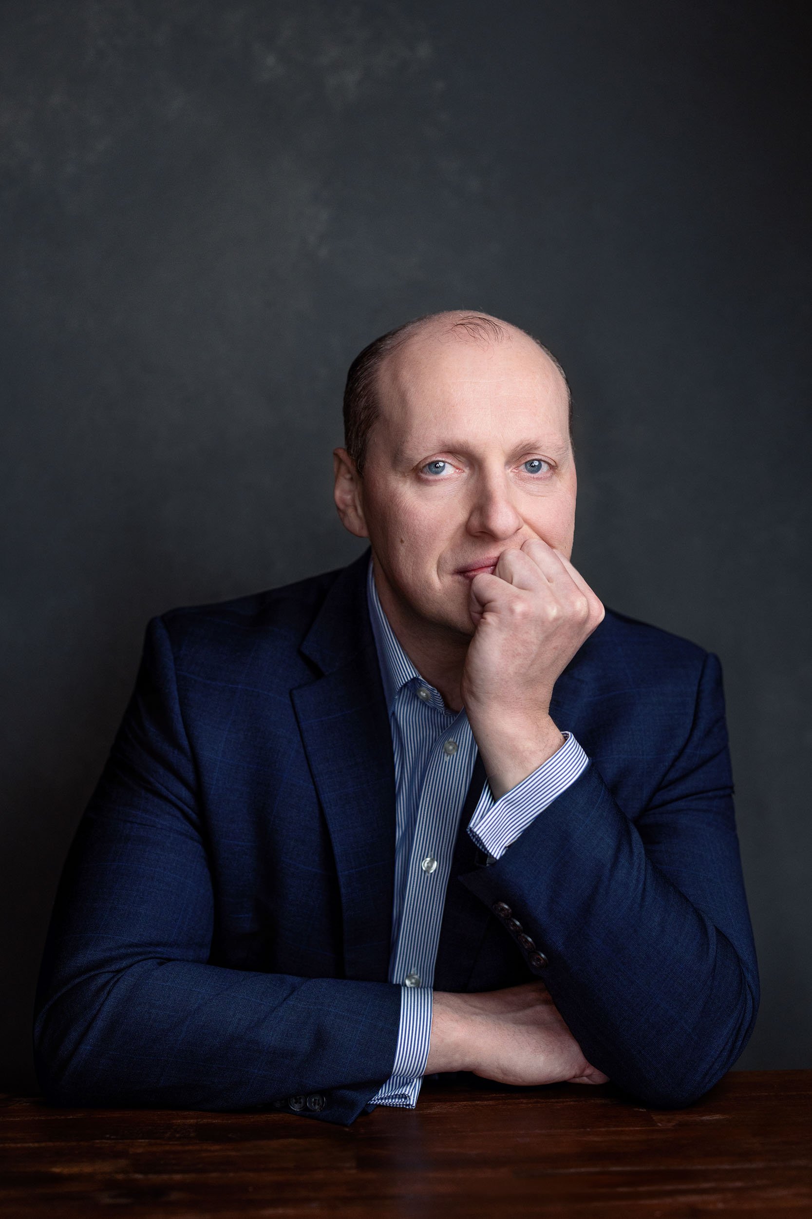 A man in a dark suit and light blue shirt sitting at a wooden table, resting his chin on his hand, looking at the camera.