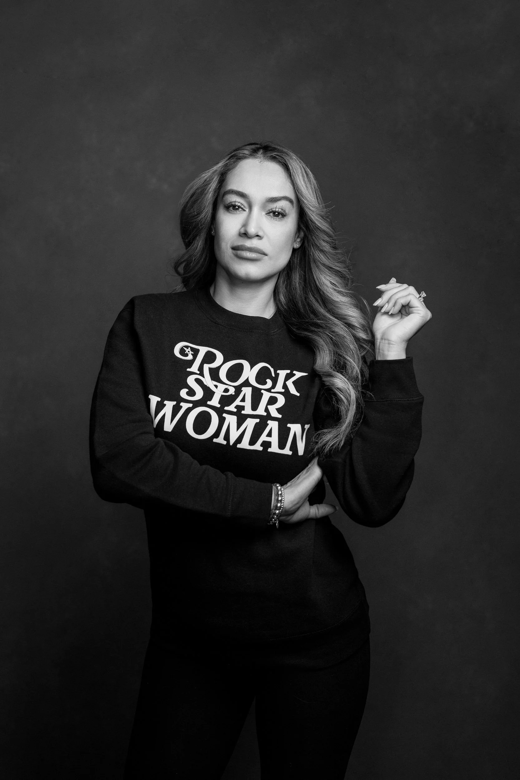 A woman with long, wavy hair wearing a black sweatshirt that reads 'Rock Star Woman', posing against a dark background in black and white.