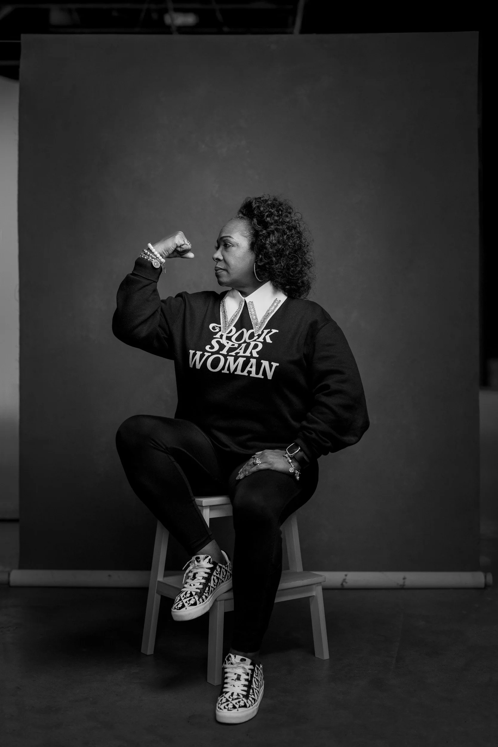 Black and white photo of a woman sitting on a stool, flexing her right arm to show her bicep, wearing a sweatshirt that says 'GIRL ROCK STAR WOMAN', with curly hair, patterned sneakers, and various bracelets and rings, against a dark background.