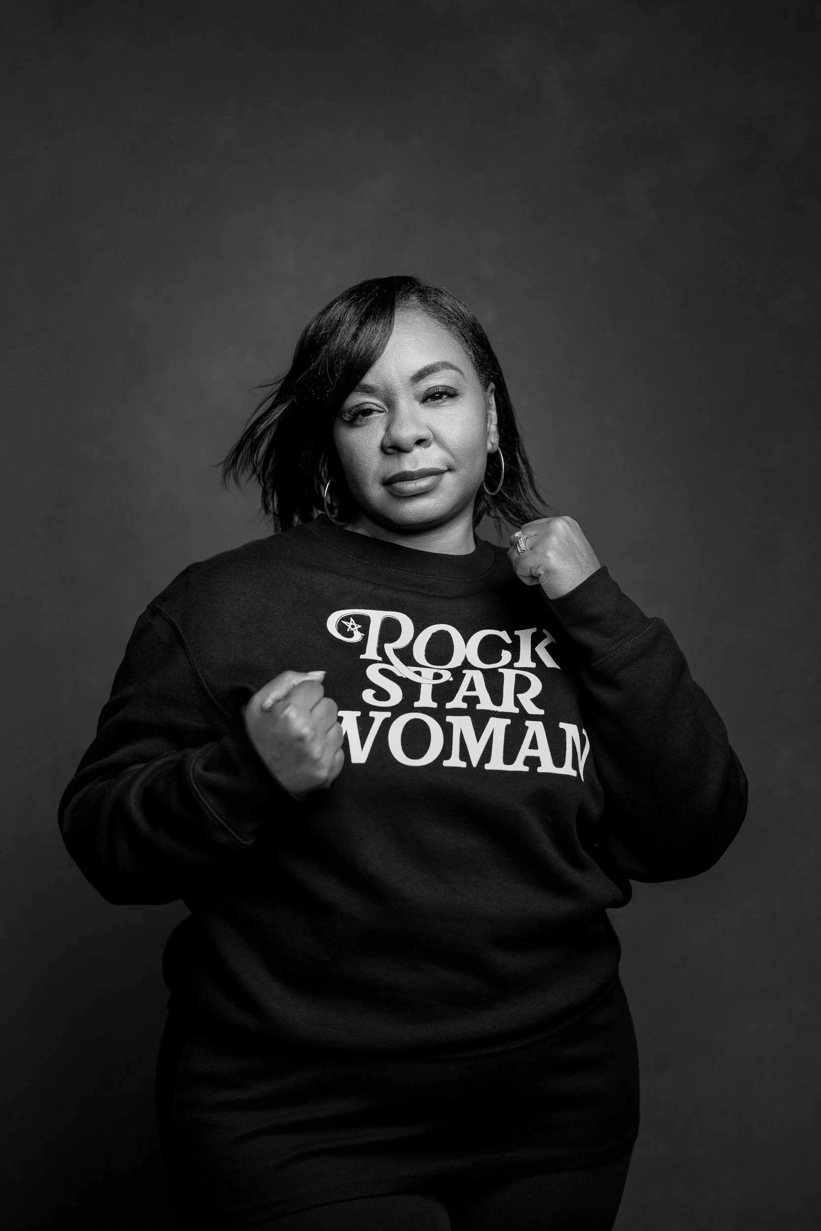 Black and white portrait of a woman with short hair, raising her fists in a confident pose, wearing a sweatshirt that reads 'Rock Star Woman'.
