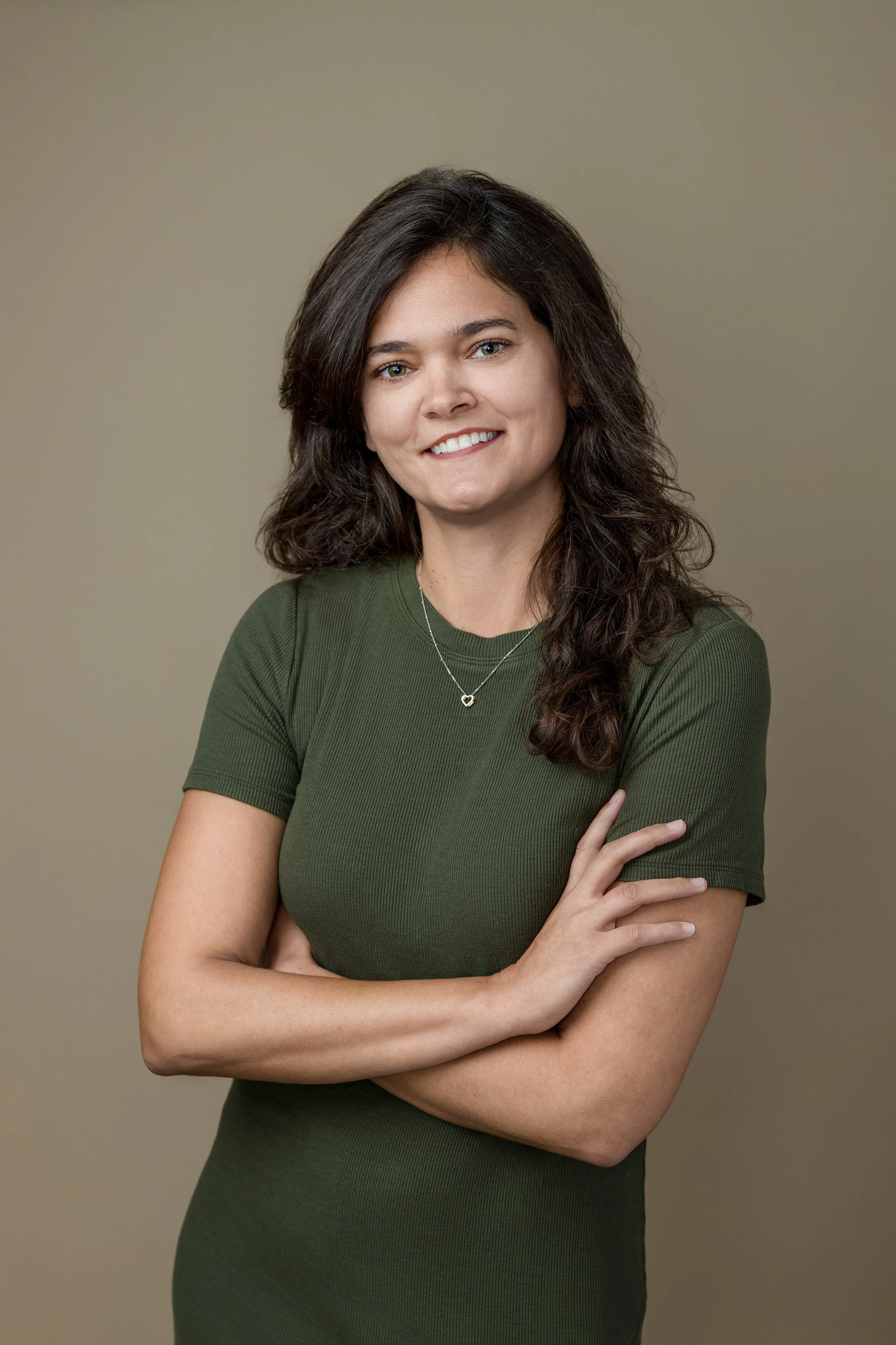 A woman with wavy brown hair smiling, wearing a green ribbed shirt and a heart-shaped pendant necklace, standing against a beige background.